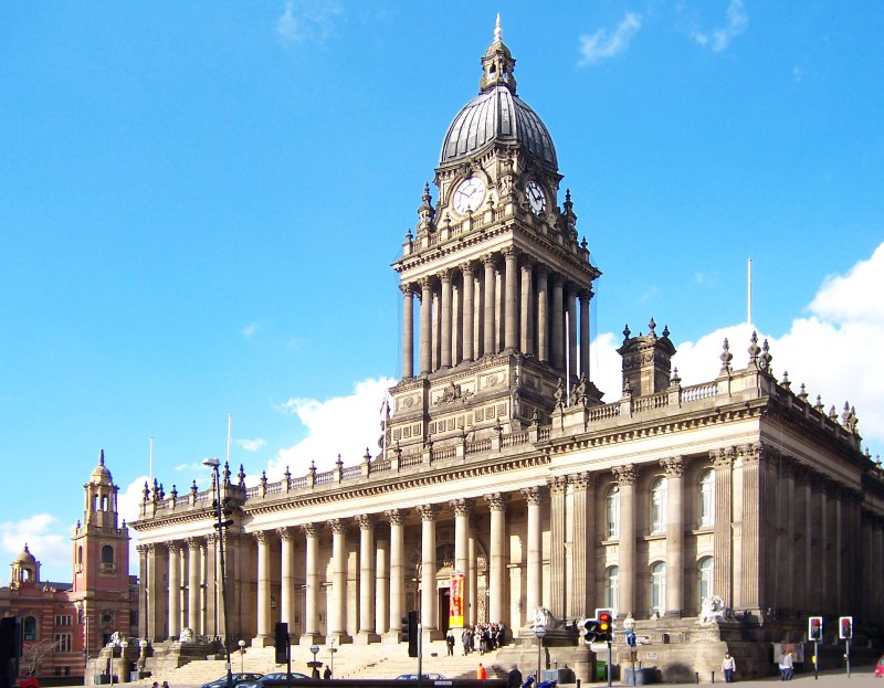A grand historic building in Leeds with a large domed clock tower and columns, set against a bright blue sky with some clouds. People are visible near the entrance, highlighting the vibrant student city atmosphere. Vehicles wait at the front.