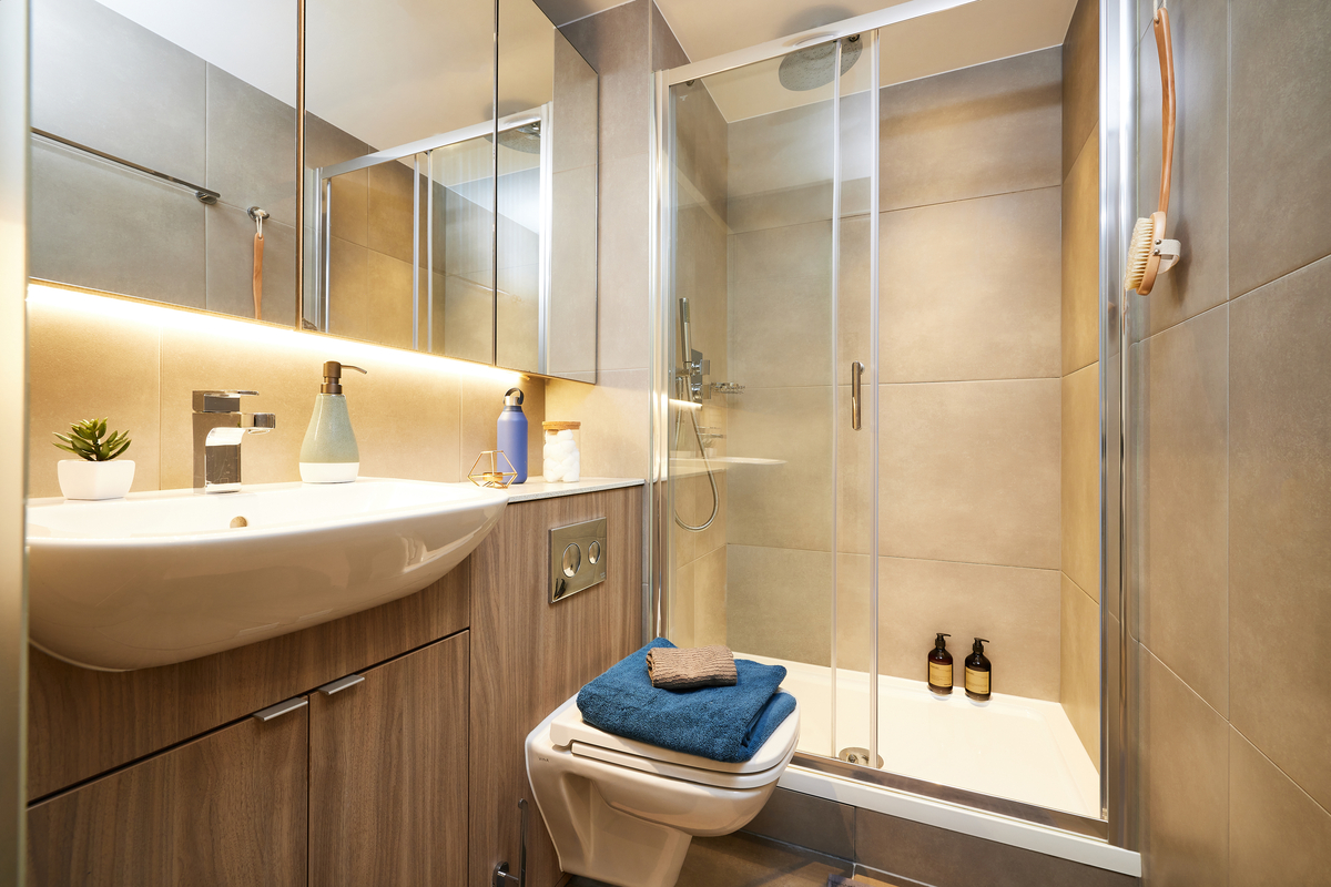 A modern bathroom in Newcastle features tiled walls, a glass-enclosed shower, and a wall-mounted sink. Blue towels and a small plant add touches of decor. Neutral tones enhance the clean, minimalist vibe.
