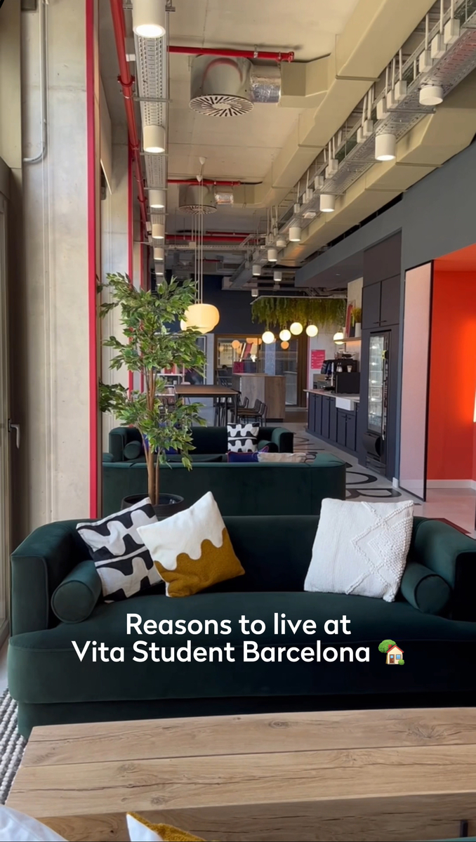 Modern lounge interior at Vita Student Barcelona, featuring green sofas with assorted cushions, a wooden table, and potted plants. The ceiling has exposed pipes and circular lights, and a wall-mounted text reads, "Reasons to live at Vita Student Barcelona.