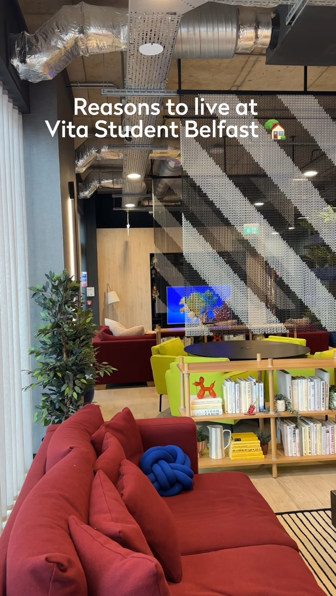 Stylish student lounge with modern red couches, a colorful bookshelf, and a wall-mounted TV. The open space features industrial decor with exposed ducts and a variety of seating options. Text overlay promotes living at Vita Student Belfast.