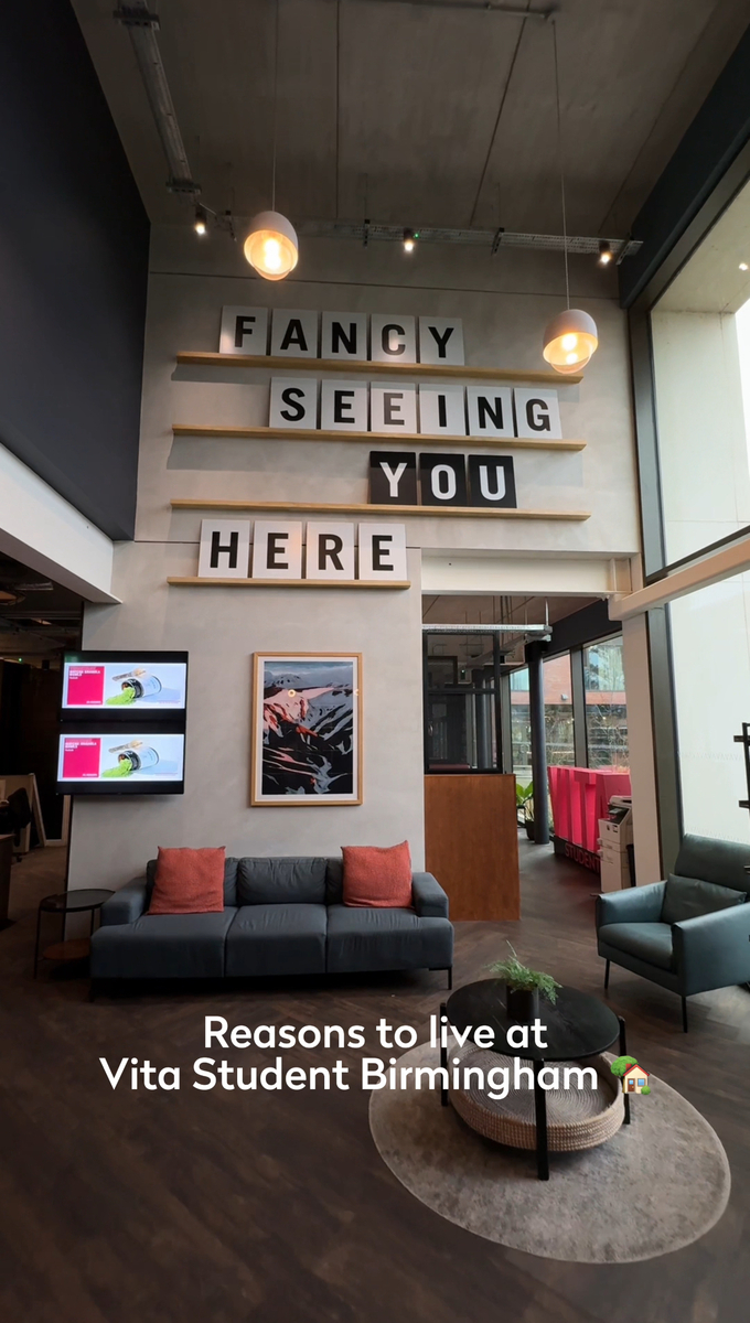 Modern lounge area with high ceiling and large windows, featuring a blue sofa, two armchairs, a coffee table with plants, and a sign saying "FANCY SEEING YOU HERE". Text overlay reads, "Reasons to live at Vita Student Birmingham.