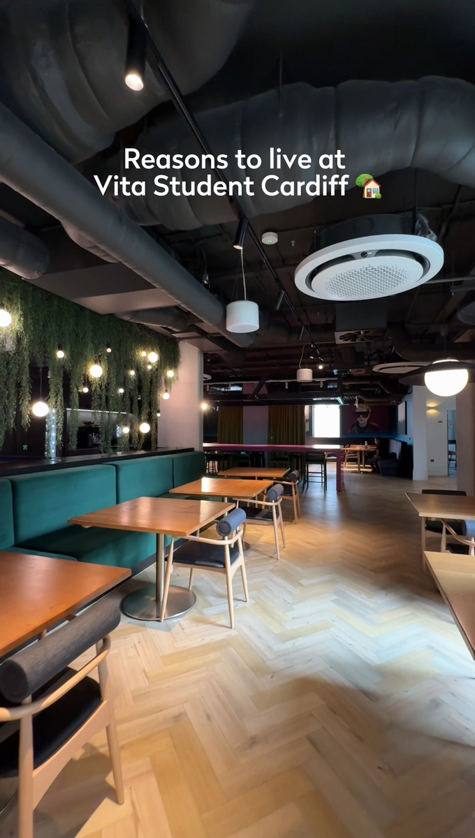 Modern student common area with wooden tables and chairs, surrounded by greenery on the walls. Overhead lighting and sleek design create an inviting atmosphere. Text reads "Reasons to live at Vita Student Cardiff.