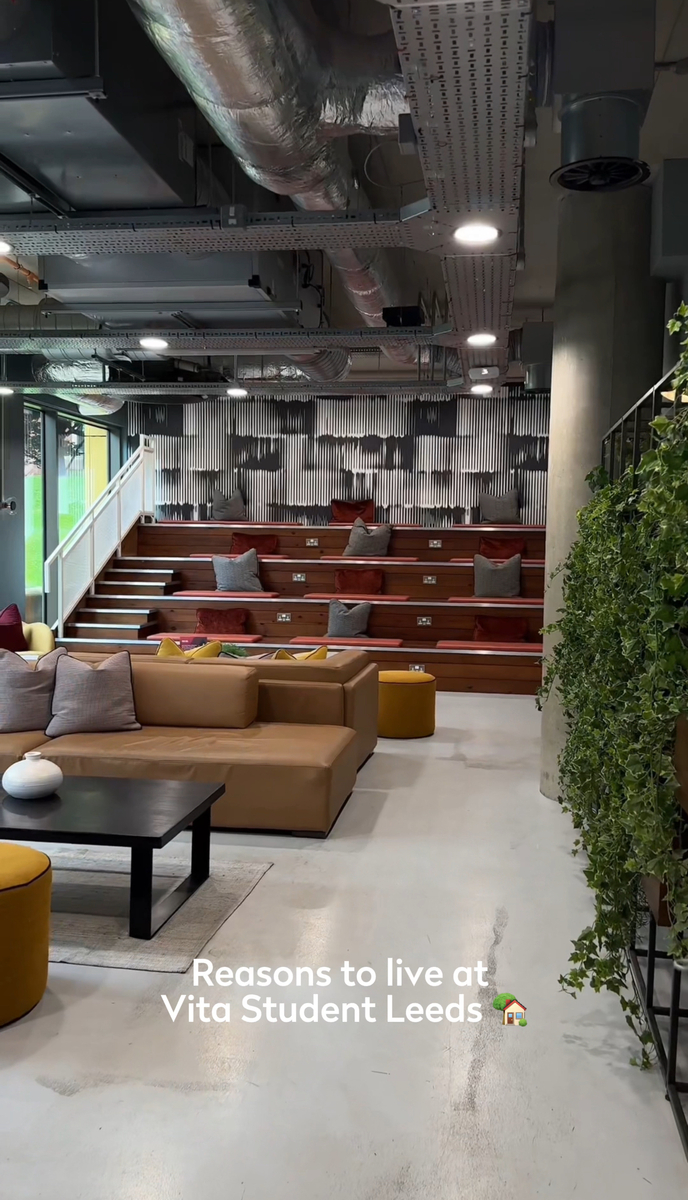 A modern lounge area with tiered seating and a row of cushions against a wall, a couch and coffee table in the foreground, and hanging plants on the side. Text reads "Reasons to live at Vita Student Leeds" with a house emoji.