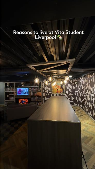 Modern interior of a communal living space at Vita Student Liverpool, featuring a long table, bookshelf, TV, and geometric patterned walls. Overhead lighting and decorative elements add a cozy atmosphere.