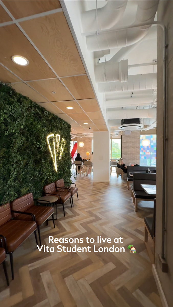A modern lounge area with a plant-covered wall and neon light, leather chairs, and tables. People are sitting in the background near large windows. Text on the image reads, "Reasons to live at Vita Student London.