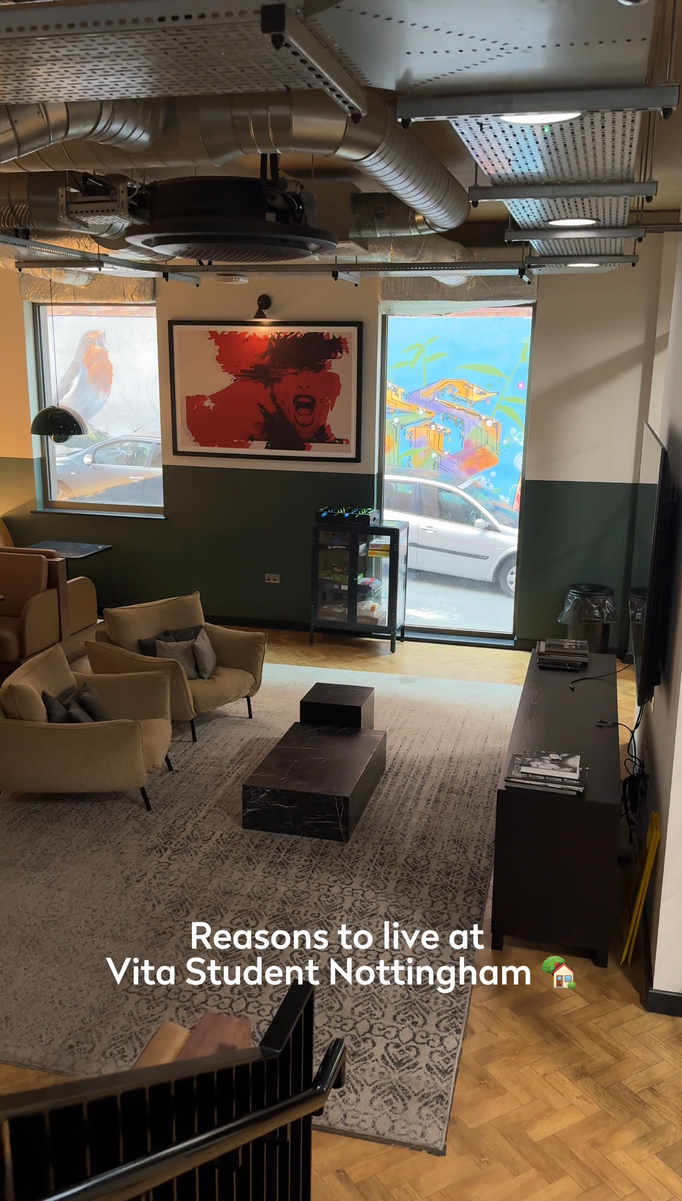 Modern lounge area with plush seating and a large wall-mounted TV. Art adorns the walls, and large windows reveal urban street views. Text reads "Reasons to live at Vita Student Nottingham.