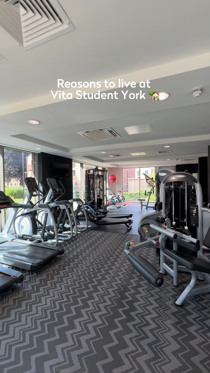 A modern gym with treadmills, exercise bikes, and weight machines. The room has large windows allowing natural light. Text reads "Reasons to live at Vita Student York" with a house emoji.