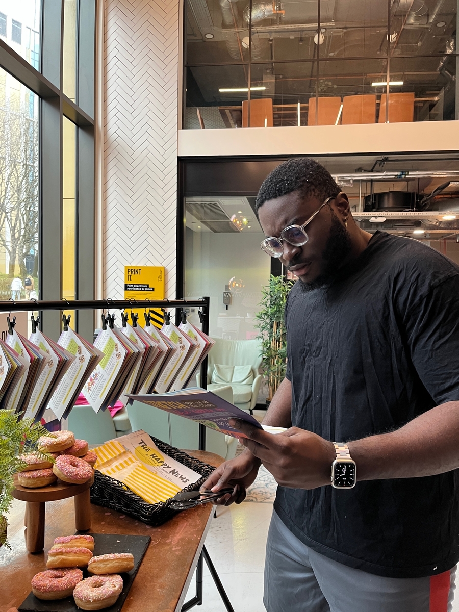 A man in a black shirt and glasses reads a menu while standing in a modern cafe. A table in front of him features a selection of donuts. Bright natural light streams in through large windows in the background.