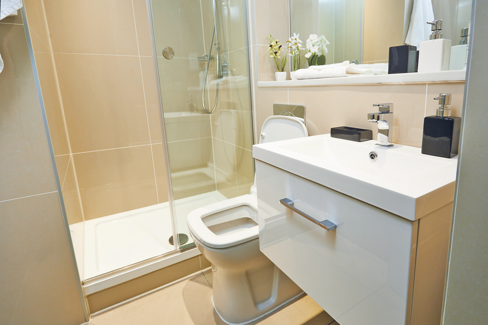 A modern bathroom featuring a glass-enclosed shower, a white toilet, and a sleek white sink with a rectangular mirror above. The counter has toiletries, a potted plant, and neatly folded towels. The walls are light beige.