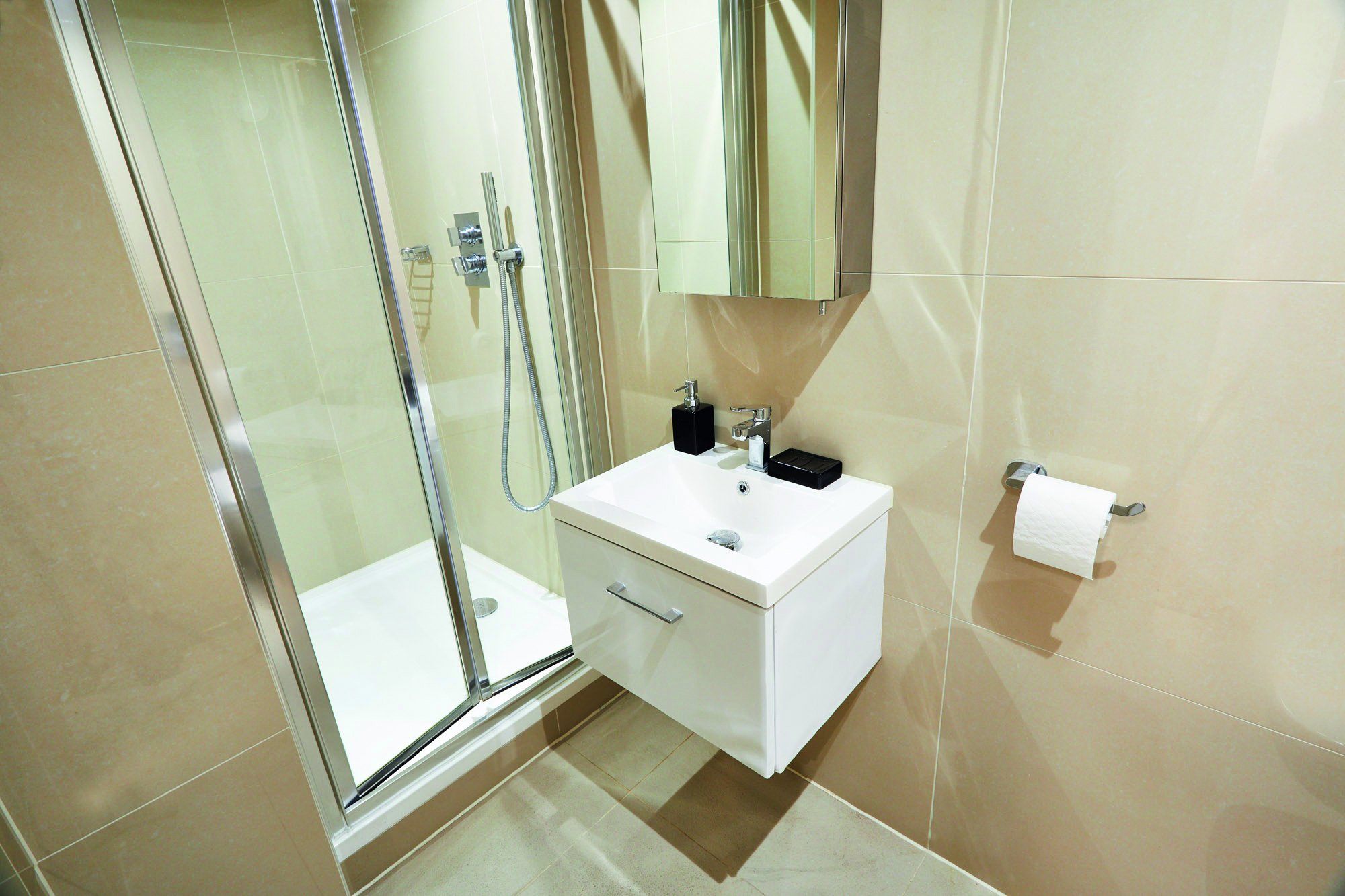 A modern bathroom in Sheffield features a glass shower enclosure with a rainfall showerhead, a sleek white sink with chrome fixtures, a mirrored cabinet above, and a toilet paper holder on the beige tiled wall.