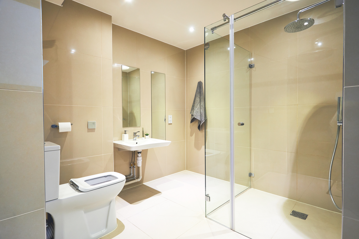 Modern bathroom with beige tiles, featuring a glass-enclosed shower and a toilet. A rectangular sink with a mirror above is mounted on the wall. A gray towel hangs nearby, and recessed lighting illuminates the space.