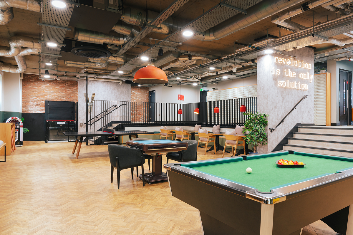 Spacious lounge area with a pool table in the foreground, a table with chairs, and a shuffleboard table. Exposed ductwork on the ceiling, industrial-style decor, and a neon sign reading "revolution is the only solution" on the wall.