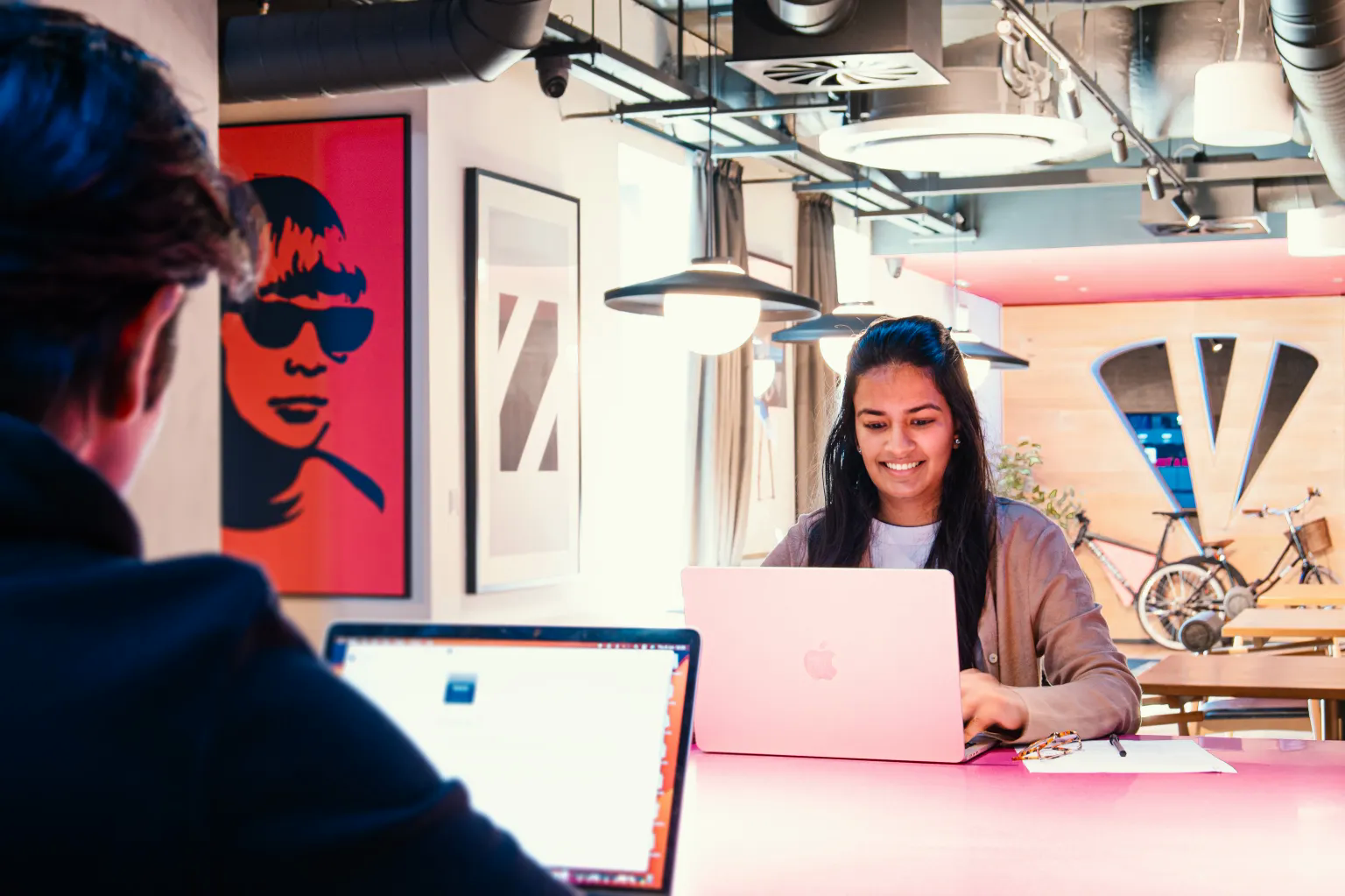 In a modern office adorned with art and a bicycle leaning nearby, two people sit across from each other at a table. The woman, smiling, navigates her laptop while the man focuses on his screen. They appear to be studying market trends in India, exchanging insights for their latest project landing.