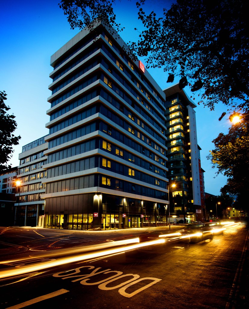 A tall, modern office building in Southampton is illuminated at dusk, with light trails from passing cars and "LOOK" painted on the road. Trees frame the top corners, highlighting a great place to study or work in the city.