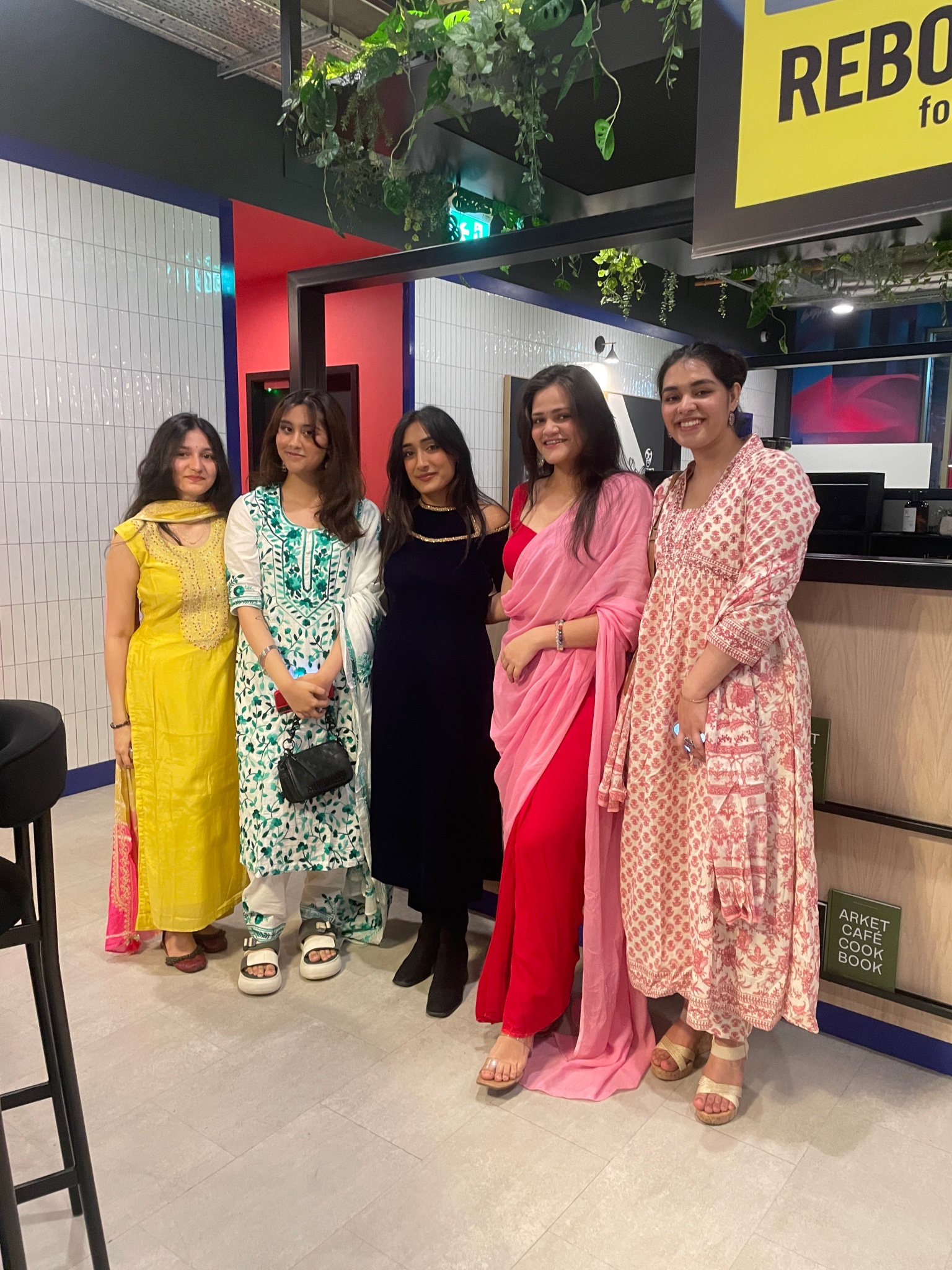 Five people standing indoors, smiling at the camera. They are dressed in colorful traditional attire, reminiscent of vibrant Indian culture, with a mix of dresses and sarees. The setting appears to be a modern, well-lit area with a tiled wall and greenery above.