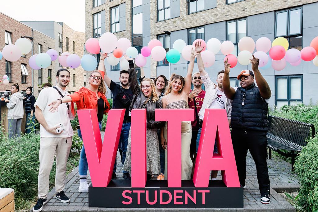 A diverse group of young adults smile and pose behind large pink “VITA STUDENT” letters in Southampton, with colorful balloons and modern apartments—showcasing a great place to live and study.