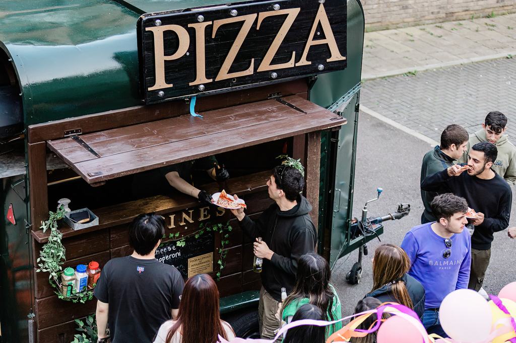 A group of people gather in front of a green pizza food truck in Southampton, with one person receiving a slice from the vendor through the window, while others stand nearby eating and chatting—it's a great place to relax after a long study session.