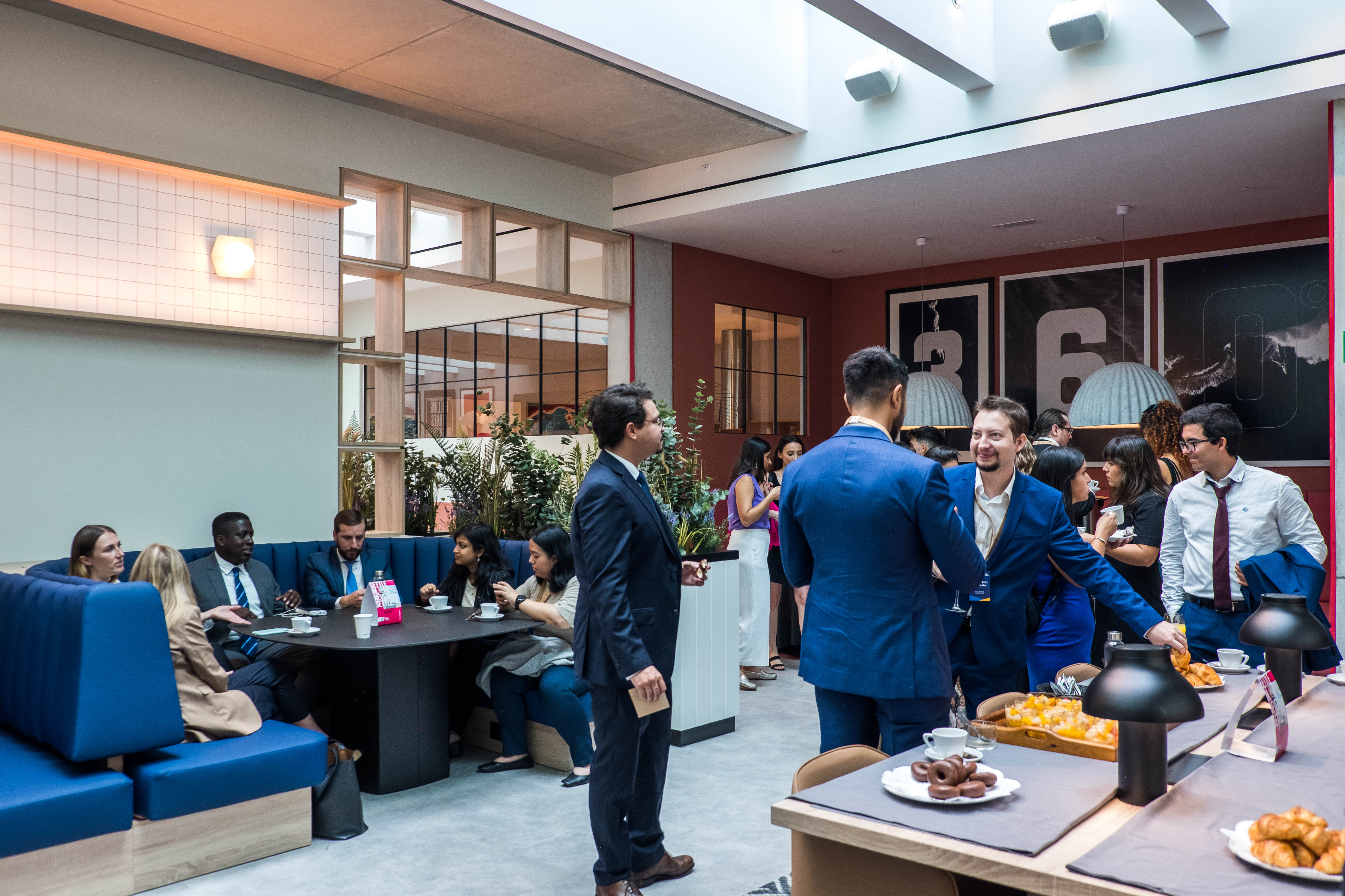 A group of people in business attire socialize, eat, and drink in a modern, brightly lit café or office lounge with large windows and a casual atmosphere—ideal for networking en España and building relaciones profesionales over pastries and drinks.