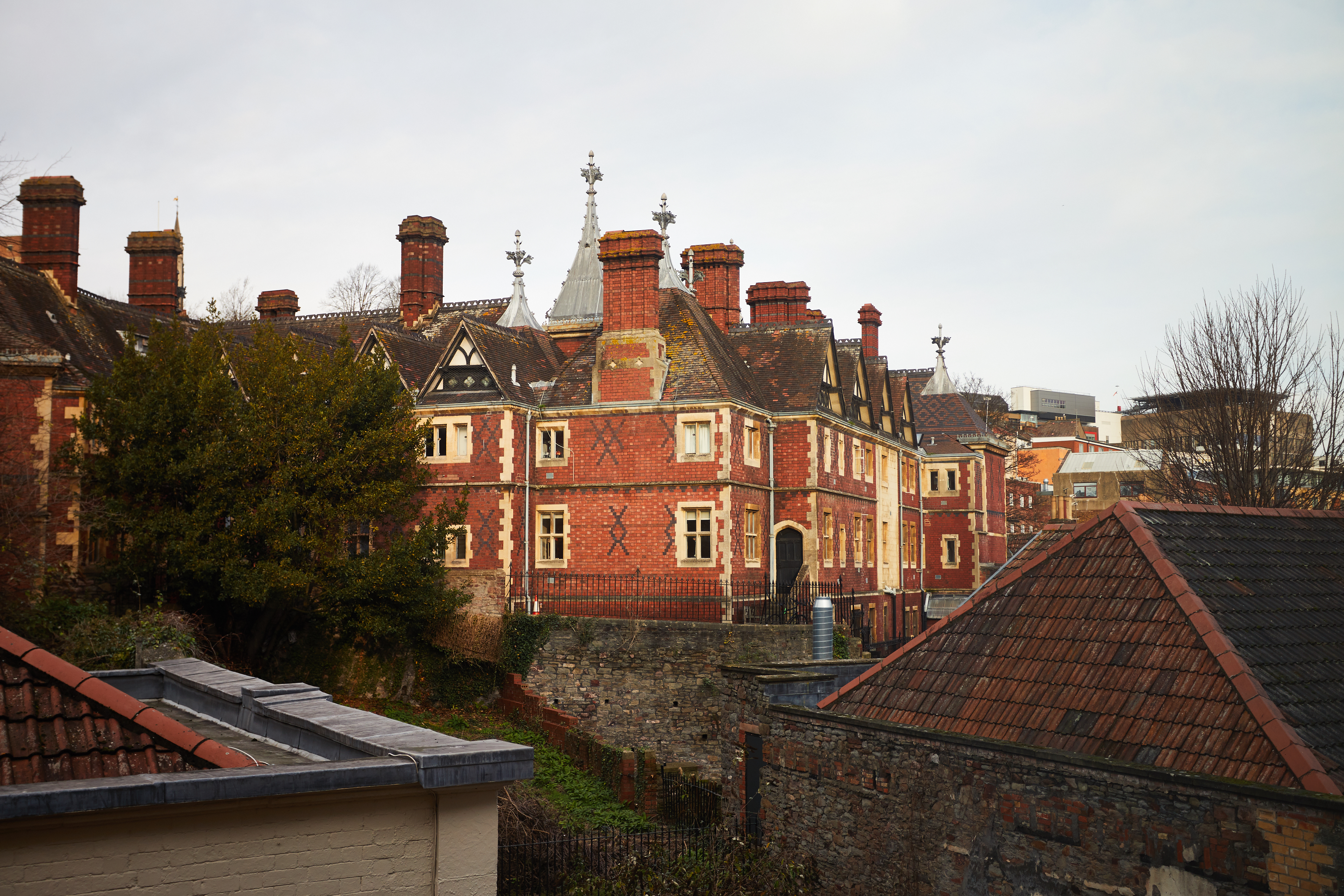 A historic red-brick building with numerous chimneys and decorative gables, often featured in films, stands surrounded by trees and rooftops under a cloudy Bristol sky.