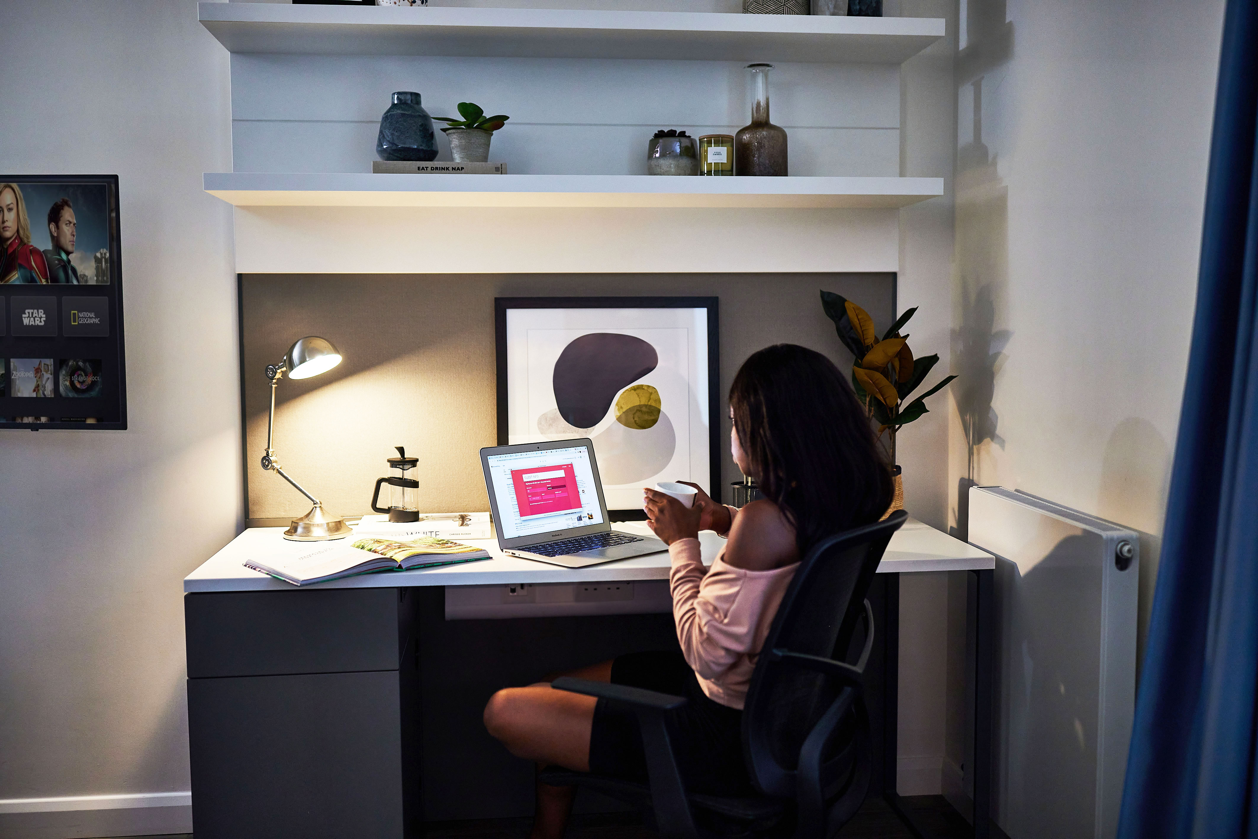 A person sits at a desk in a cozy, modern room, looking at their phone while exploring the benefits of booking online. A laptop with a bright screen is open in front of them, with shelves and a small plant visible in the background.