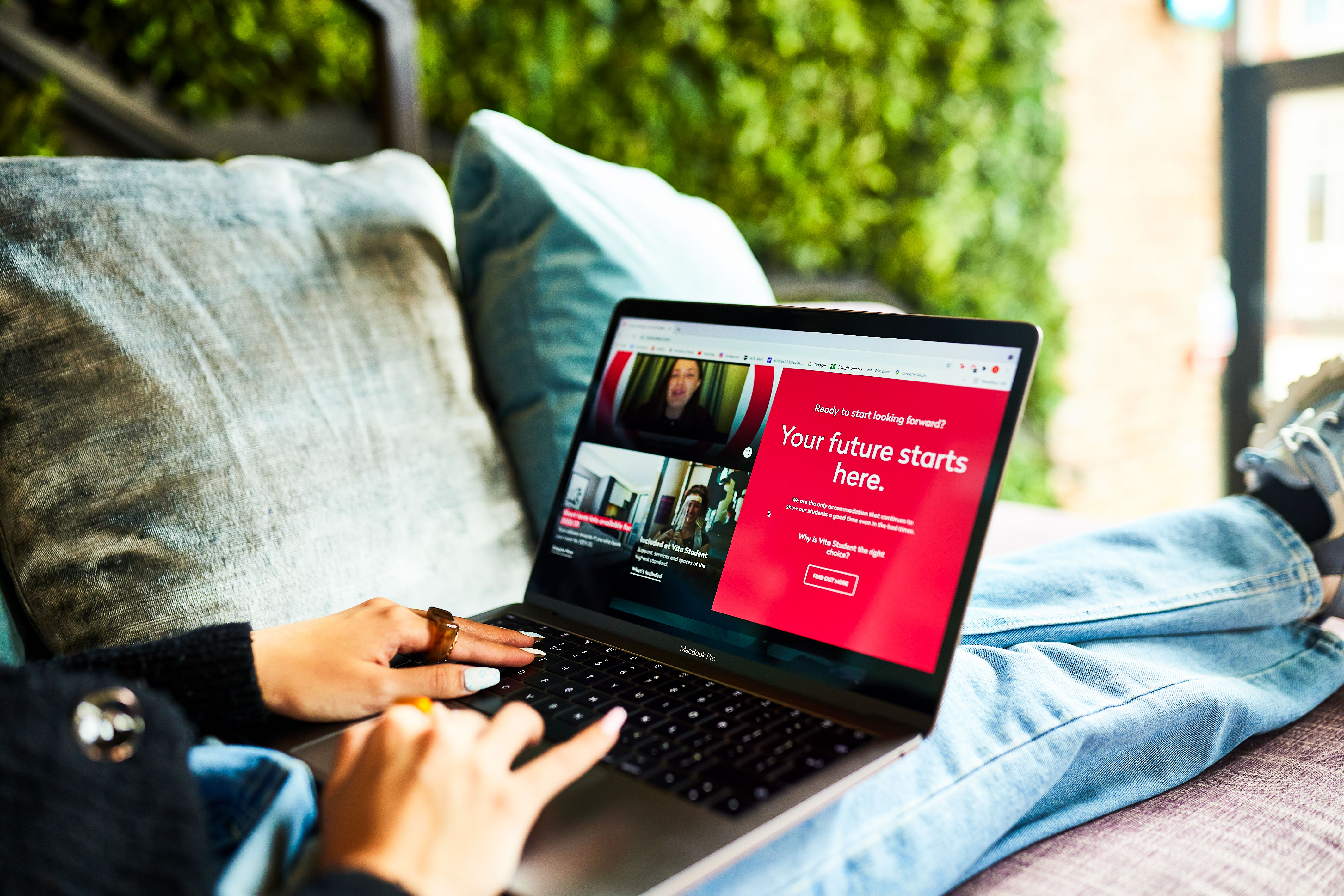 A person sits on a couch typing on a laptop. The screen displays a red Vita webpage with the text "Your future starts here." The person wears jeans and a black sweater, with a green wall in the background. Online booking benefits await.