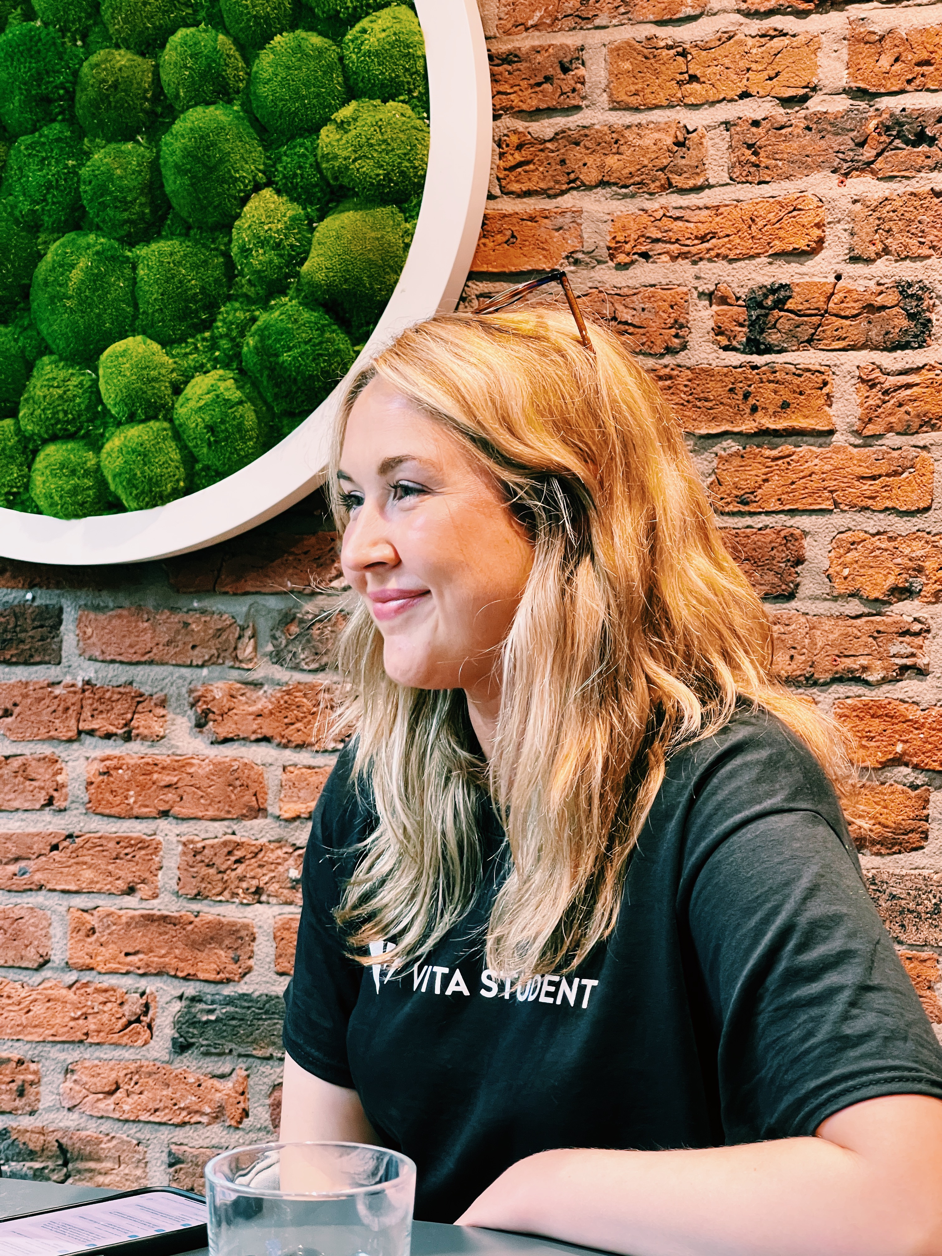 A young woman with long blonde hair sits at a table in front of a brick wall with a circular green moss decoration. She smiles gently in her dark "VITA STUDENT" t-shirt, ready to share the benefits of online booking. A glass of water is on the table.