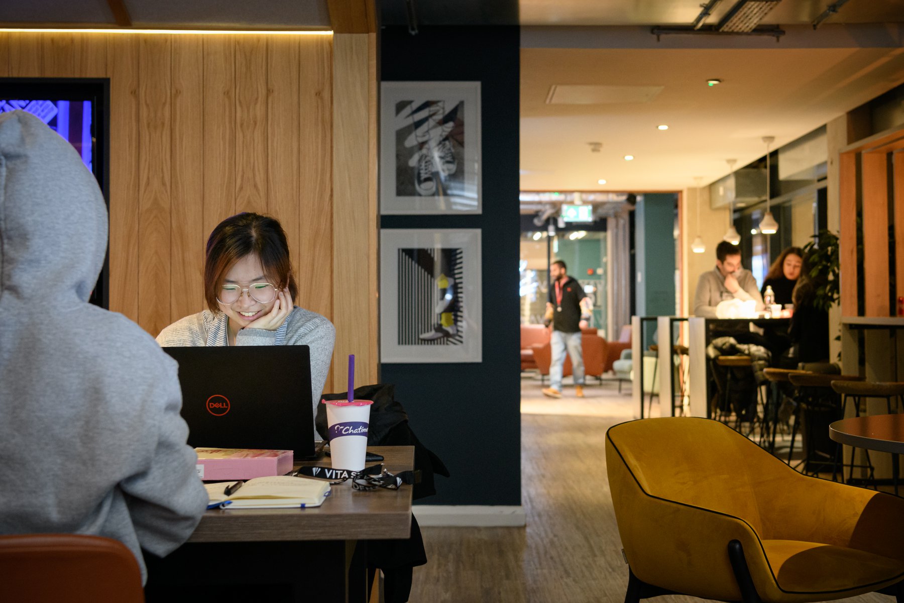 Two people sit at a table working on laptops in a cozy, modern café. One person smiles at their screen, enjoying the online benefits of Vita. Others chat and walk in the background. Books and drinks are on the table. Warm lighting fills the space.