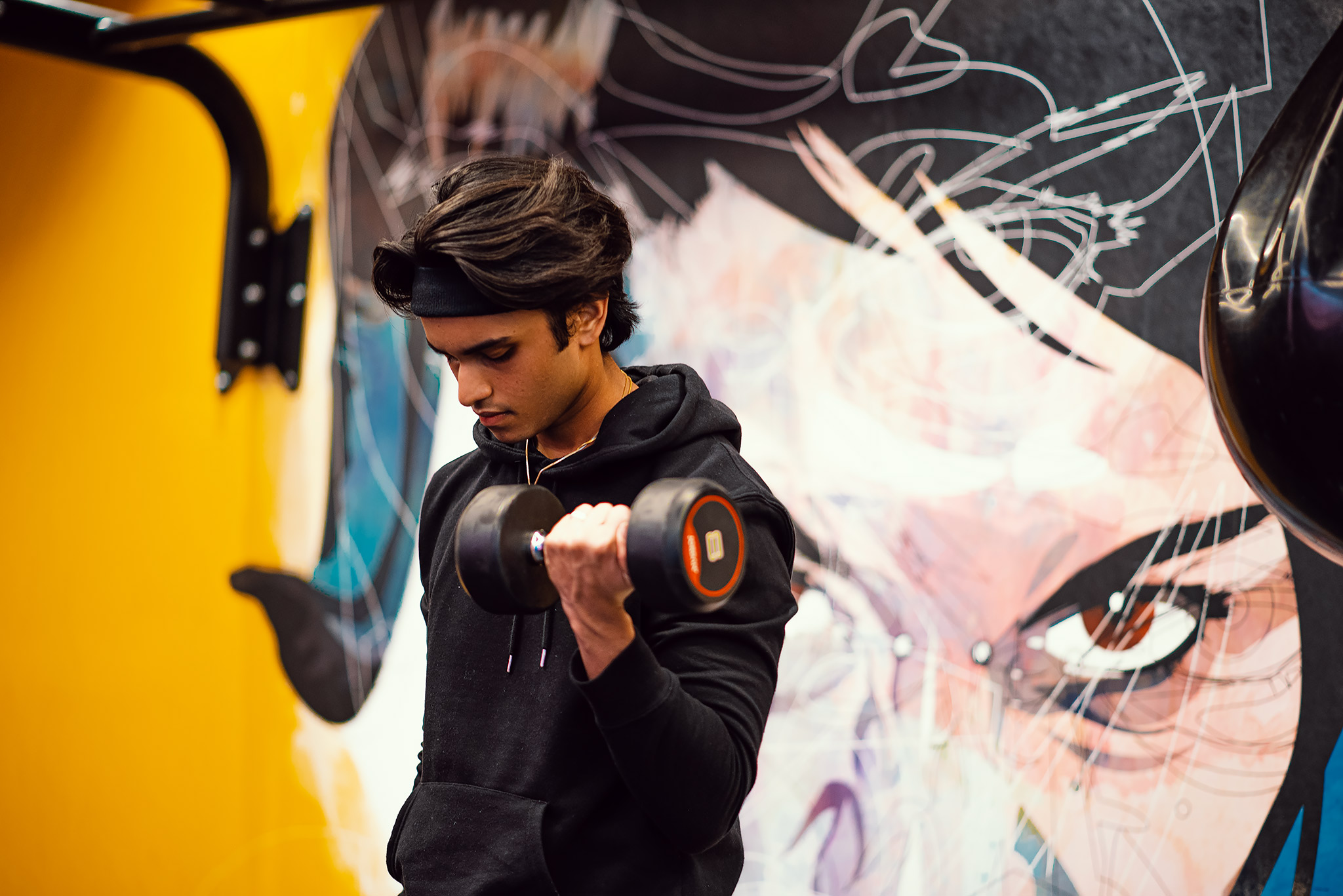 A young man in a black hoodie lifts a dumbbell in a Vita gym, standing before a colorful wall mural with abstract art and striking eyes—sign up by booking online for your own inspiring workout.