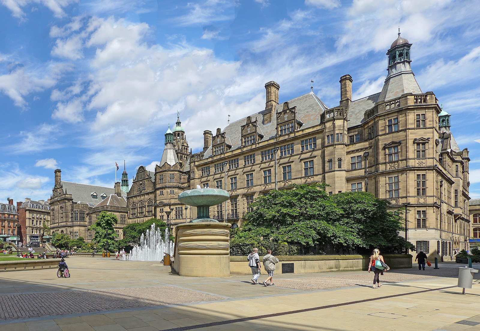 A large, ornate stone building with towers and arched windows stands behind a fountain and green trees in Sheffield, while people walk across a paved square on a sunny day with scattered clouds.