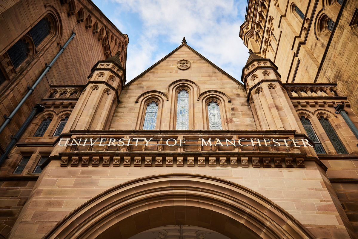 A low-angle view of the main entrance to the University of Manchester, a prestigious institution in the QS World University Rankings 2026, featuring historic stone architecture, arched windows, and the university name displayed above the doorway.