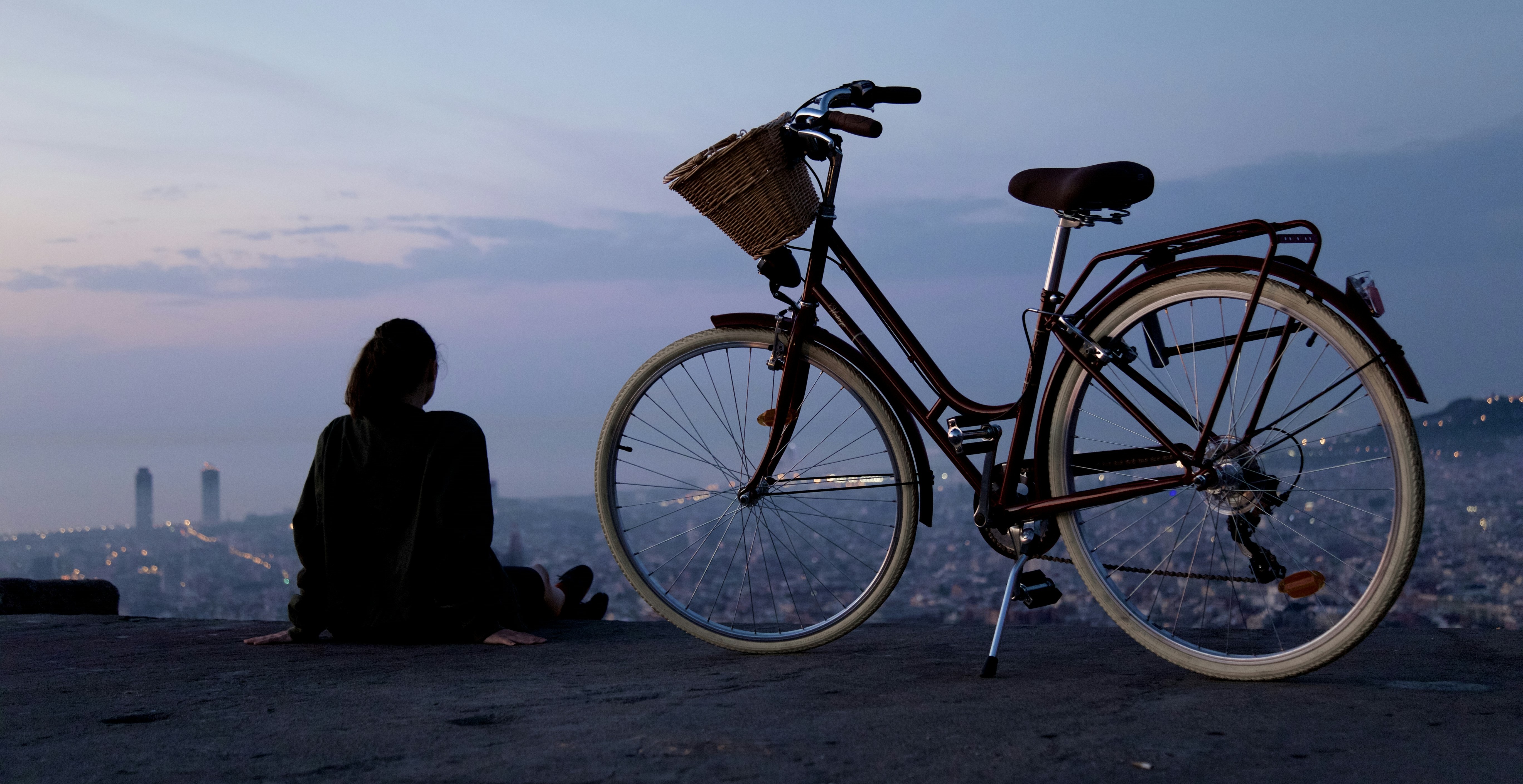 A person sits on a hill at dusk, overlooking Vita Student Barcelona and the cityscape with two tall buildings in the distance. Their bicycle stands nearby, perfect for exploring rutas en bicicleta as city lights twinkle under the blue evening sky.