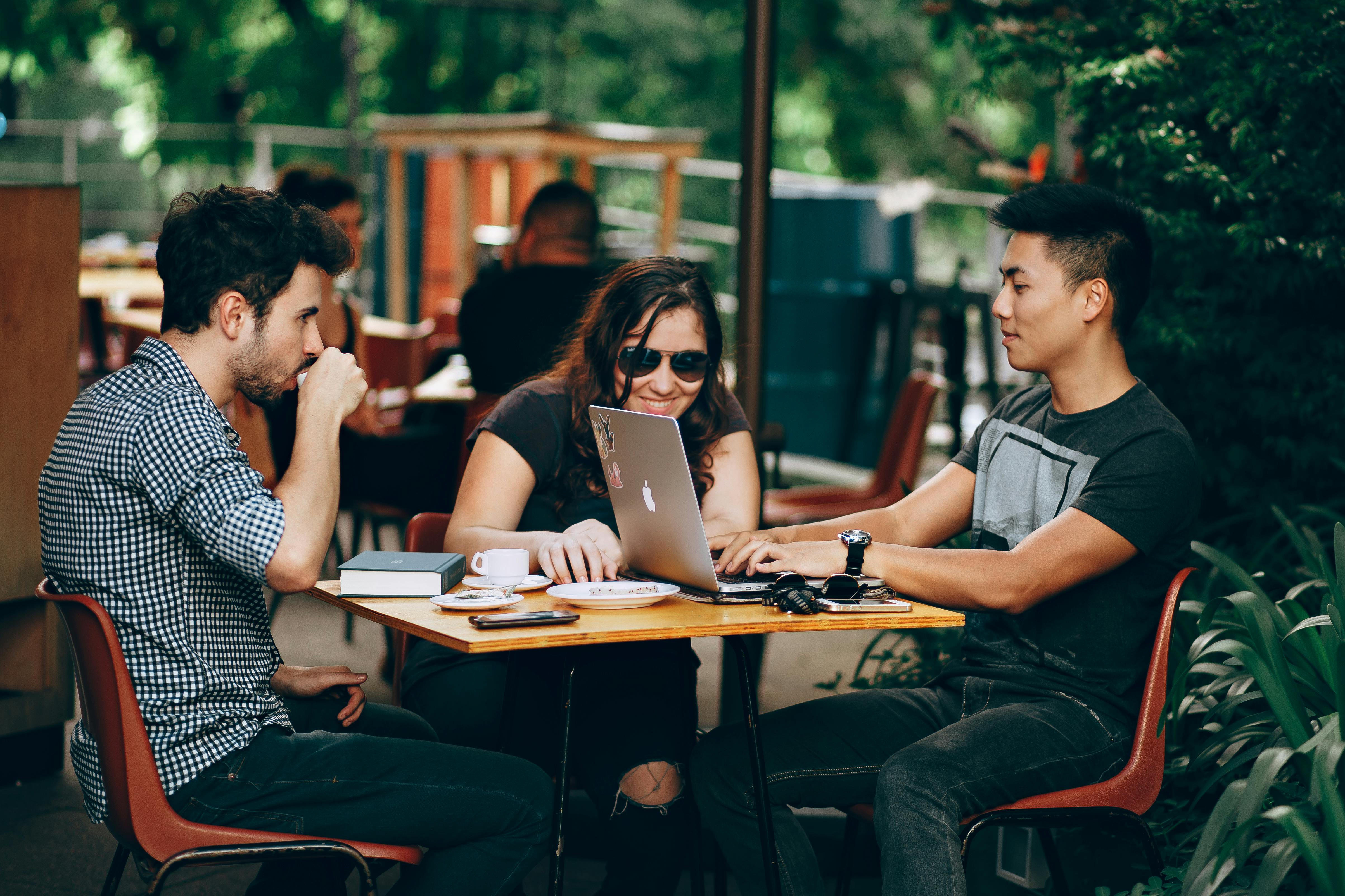 Three people sit at an outdoor table with drinks, two working on a laptop and one drinking from a cup. They appear to be collaborating or socializing as Nómadas Digitales in a casual, green, and sunny setting in España.