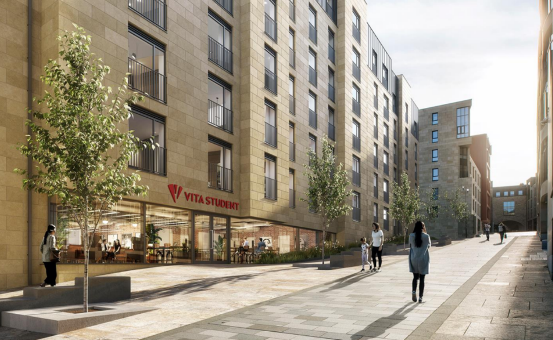 A modern student accommodation building with large windows, labeled "Vita Student," lines a wide pedestrian walkway with young trees and people walking in front of it on a sunny day.