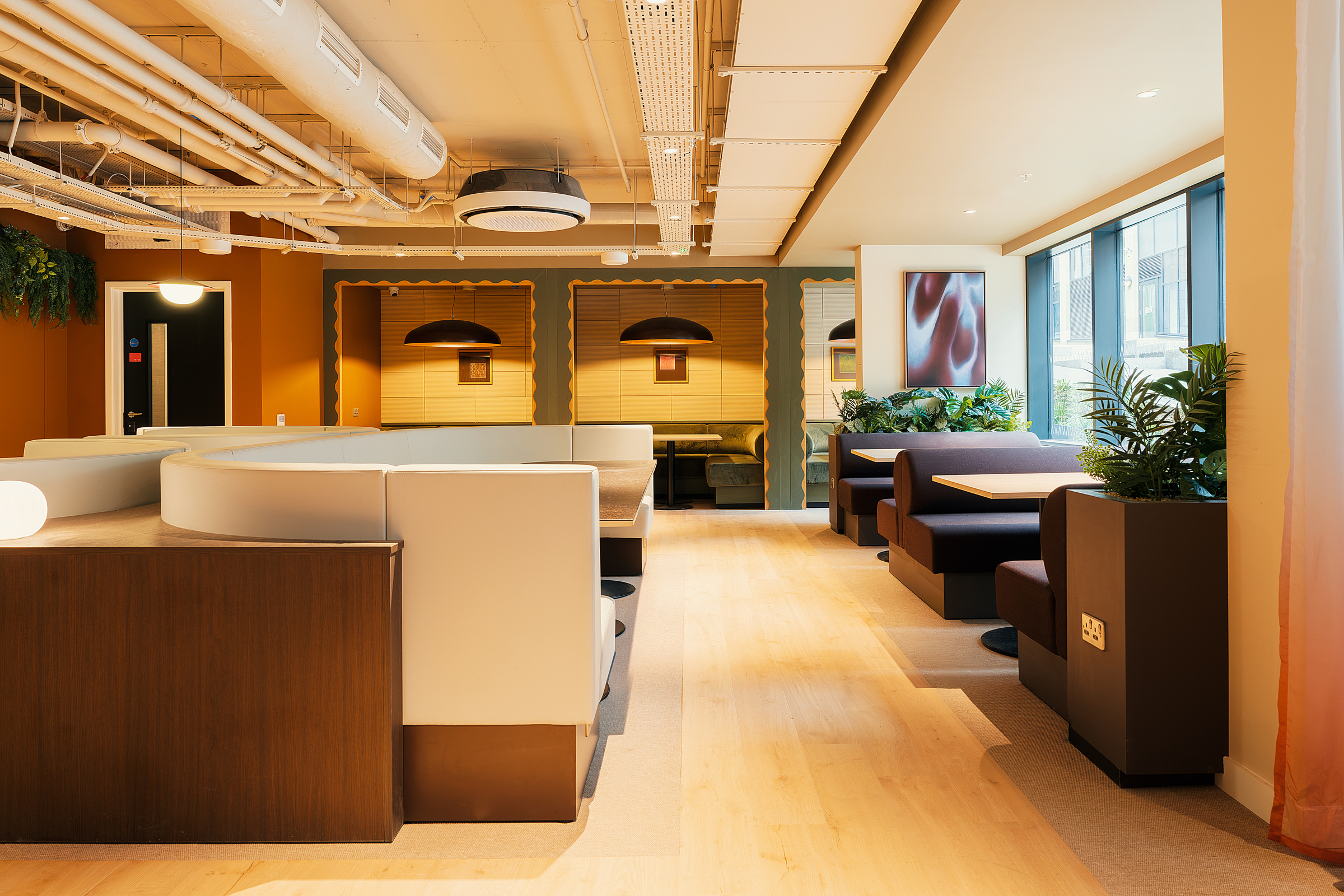 Modern office lounge with booths, light wood floors, exposed ceiling pipes, plants, and large windows letting in natural light. Cream, brown, and green tones create a cozy, contemporary atmosphere—reflecting the vibrant spirit of New Waverley.
