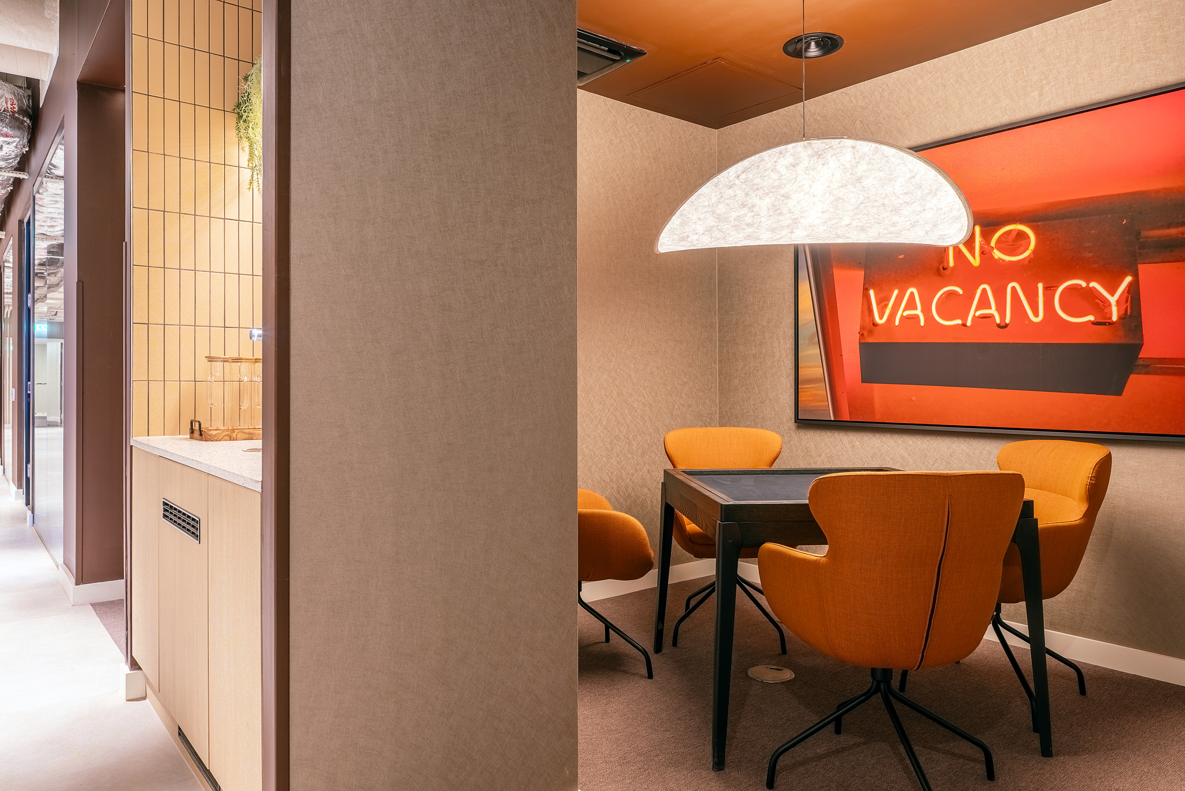 A modern meeting nook at New Waverley features four orange chairs, a dark table, a large curved pendant lamp, and a “NO VACANCY” neon sign on the wall. Neutral-toned walls and a kitchenette are visible to the left.