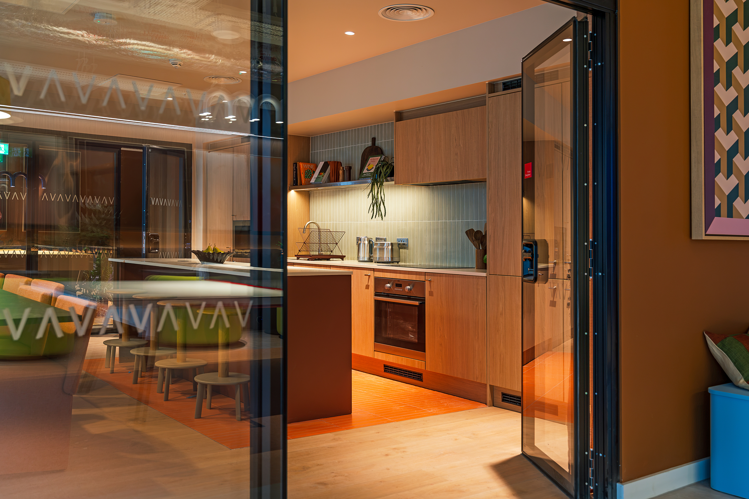 Modern kitchen with wood cabinets, an oven, and shelves is seen through a glass door in the stylish New Waverley development, next to a dining area with round tables and stools. Warm lighting creates a cozy atmosphere.