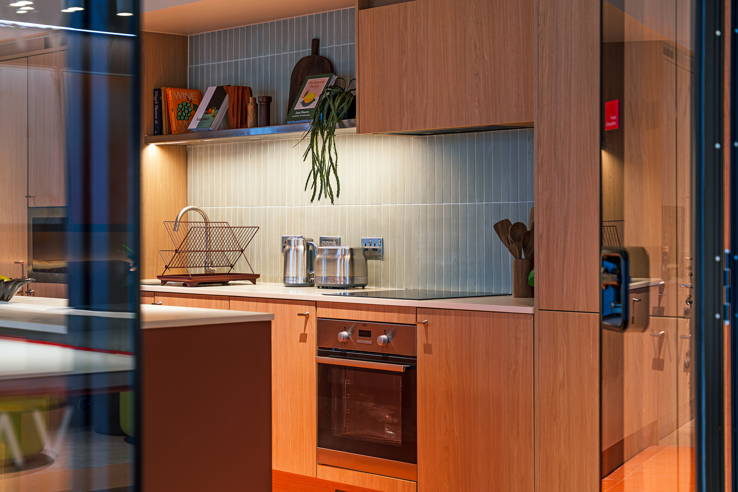 Modern kitchen with wooden cabinets, built-in oven, countertop appliances, a dish rack, cookbooks, and a plant on a shelf. Warm lighting highlights the clean, organized space behind a glass door—perfect for stylish living in New Waverley.