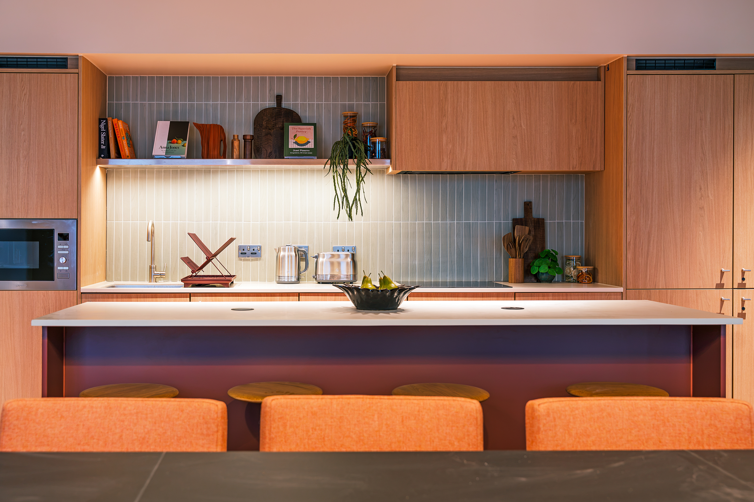 Modern kitchen in New Waverley with wooden cabinets, a white countertop, orange bar stools, small appliances, and decorative items on shelves. A fruit bowl sits on the island while warm lighting creates a cozy atmosphere.