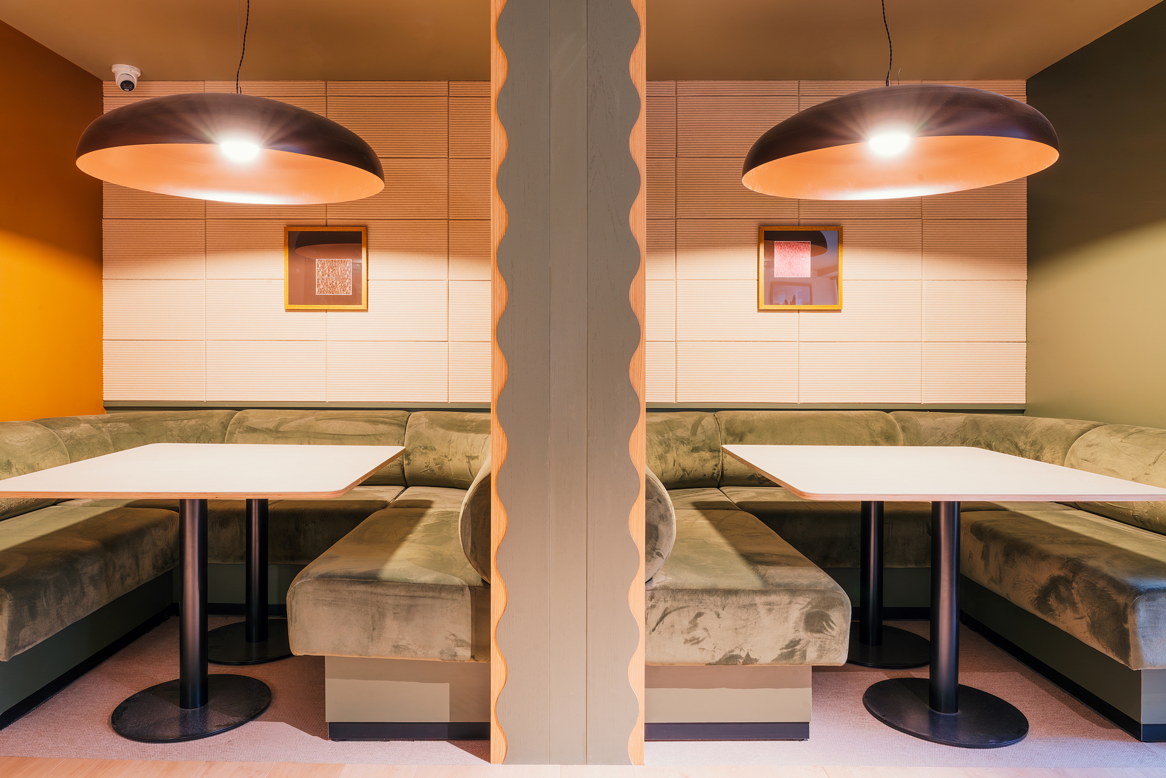 Two green velvet booth seating areas are divided by a central wavy partition, each with a white table, black pedestal base, pendant light above, and framed art on a beige wall. The space at New Waverley boasts a modern, cozy design.