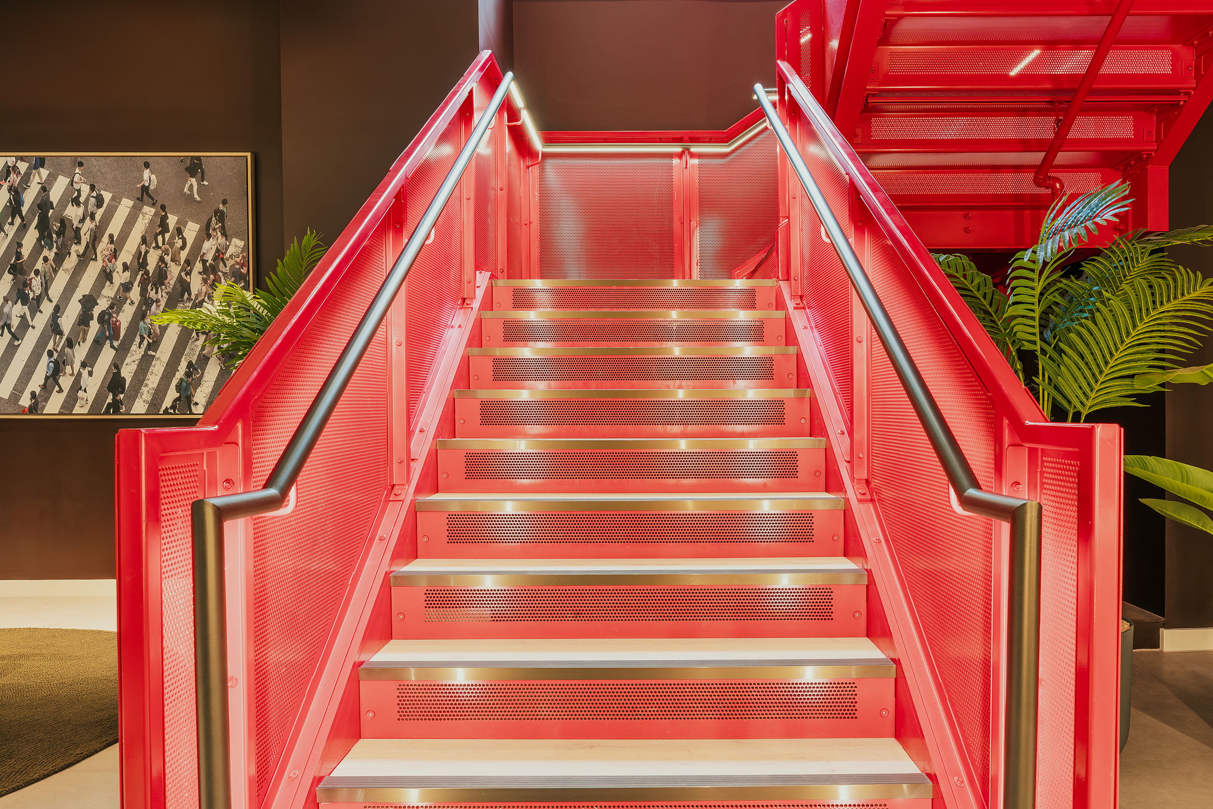 Bright red metal staircase with illuminated steps, flanked by plants, in the vibrant heart of New Waverley. A photograph of a crosswalk with people is visible on the wall to the left of the stairs.