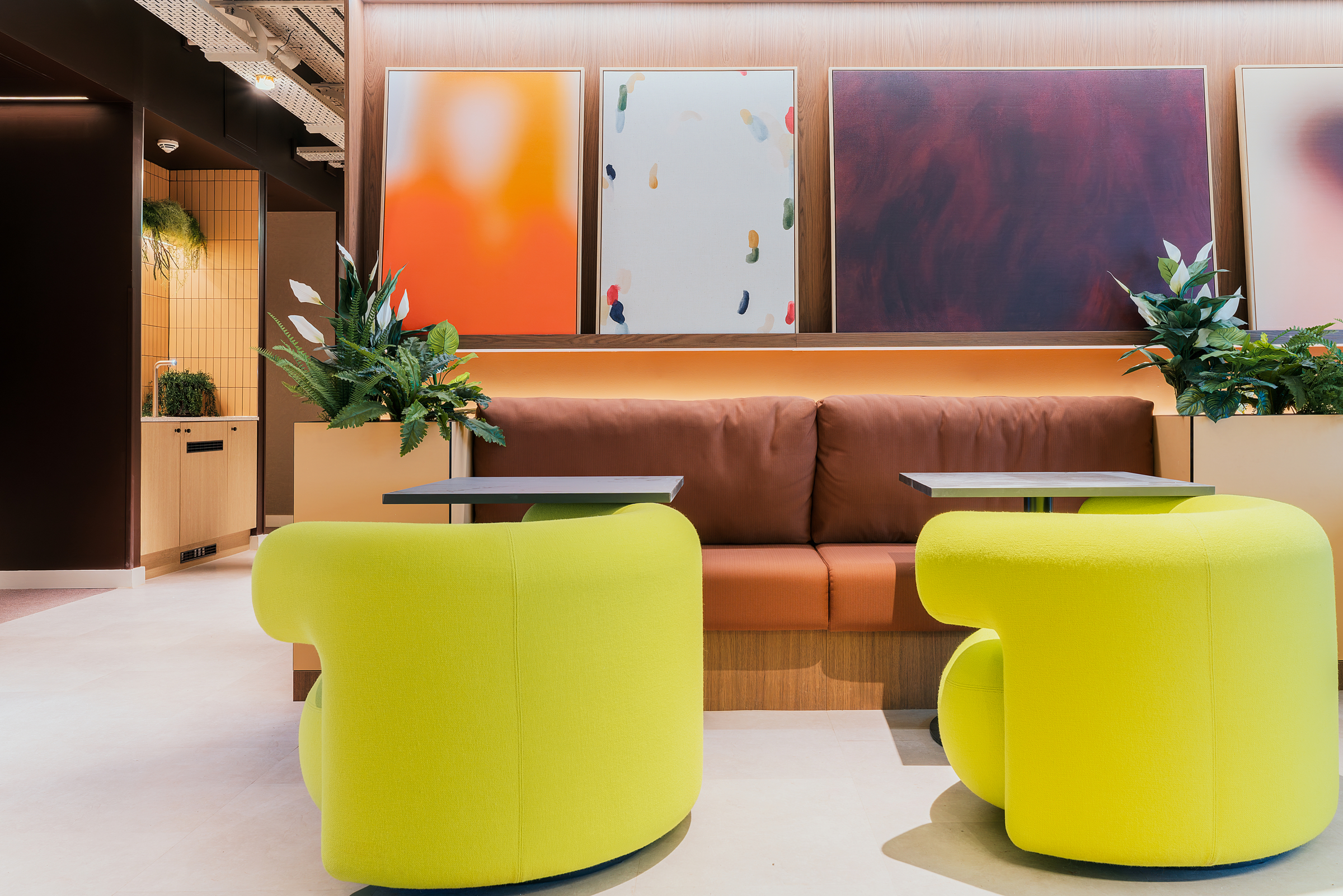 Modern lounge area with two bright yellow chairs, a brown cushioned bench, two small tables, abstract art on the wall, and green potted plants on either side—perfect for relaxing or socializing in the heart of New Waverley.