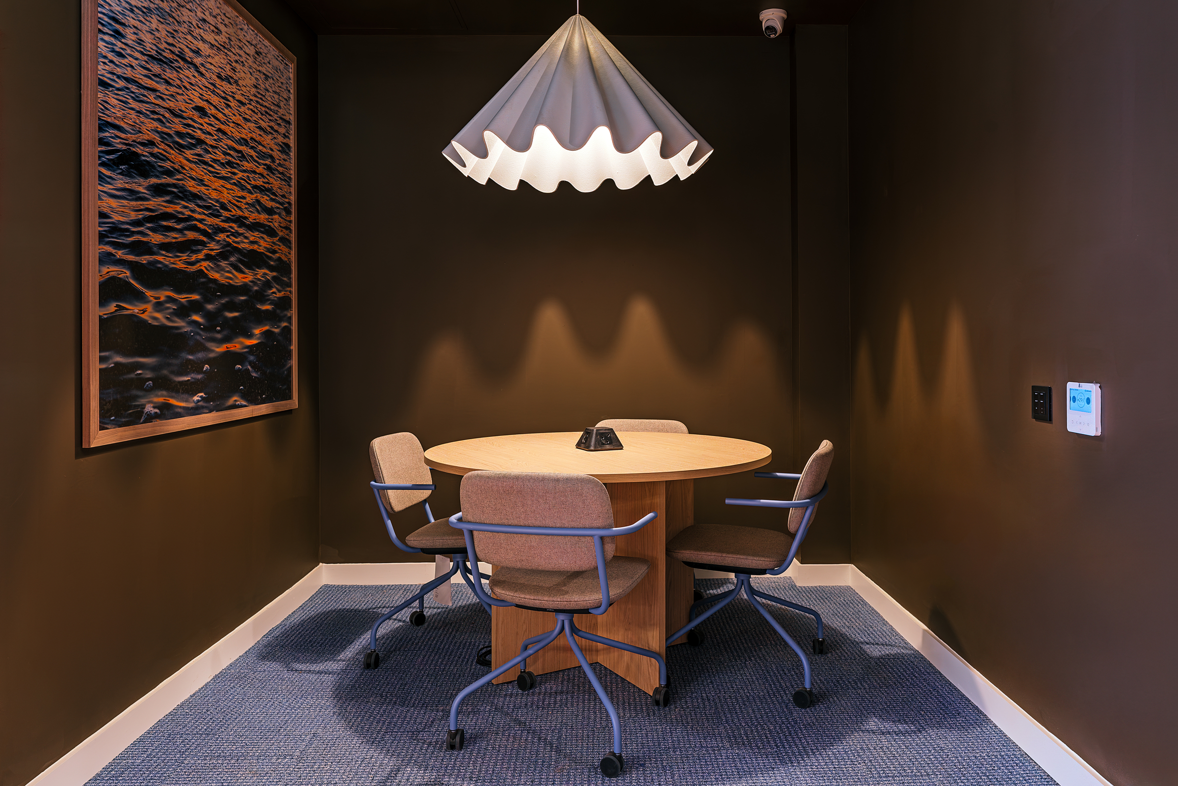 A small meeting room at New Waverley features a round wooden table, four beige chairs on wheels, a modern pendant light, a large framed picture of water, and dark brown walls complemented by blue carpeted flooring.