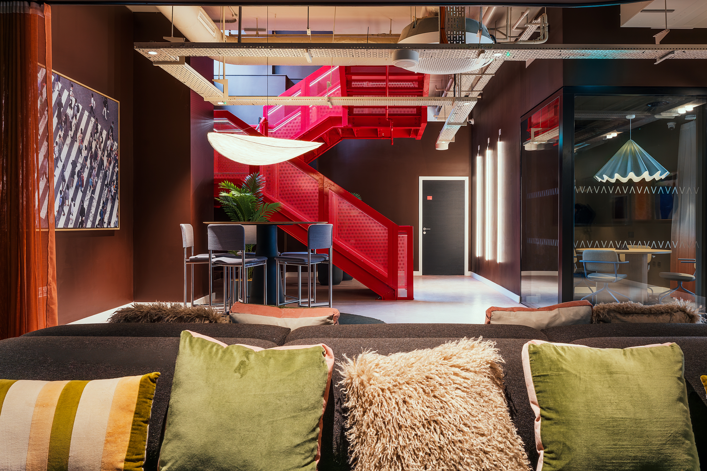 A modern lounge at New Waverley features plush pillows on a sofa, a table with chairs, plants, and a bold red staircase in the background. Artwork and a unique ceiling light add style to the contemporary interior.