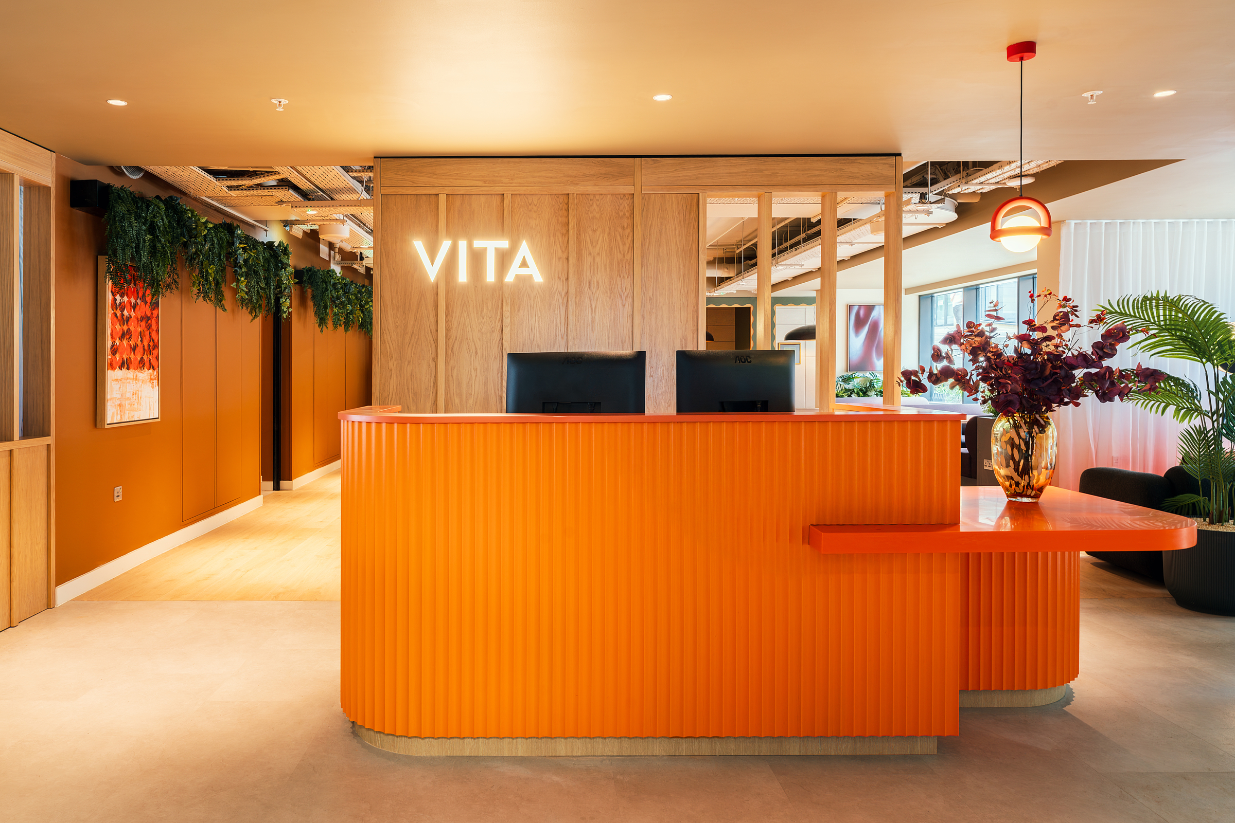 A modern reception area with an orange curved desk, two monitors, a vase with flowers, and “VITA” illuminated on a wood panel wall. Inspired by the vibrant energy of New Waverley, the space features plants, warm lighting, and contemporary decor.