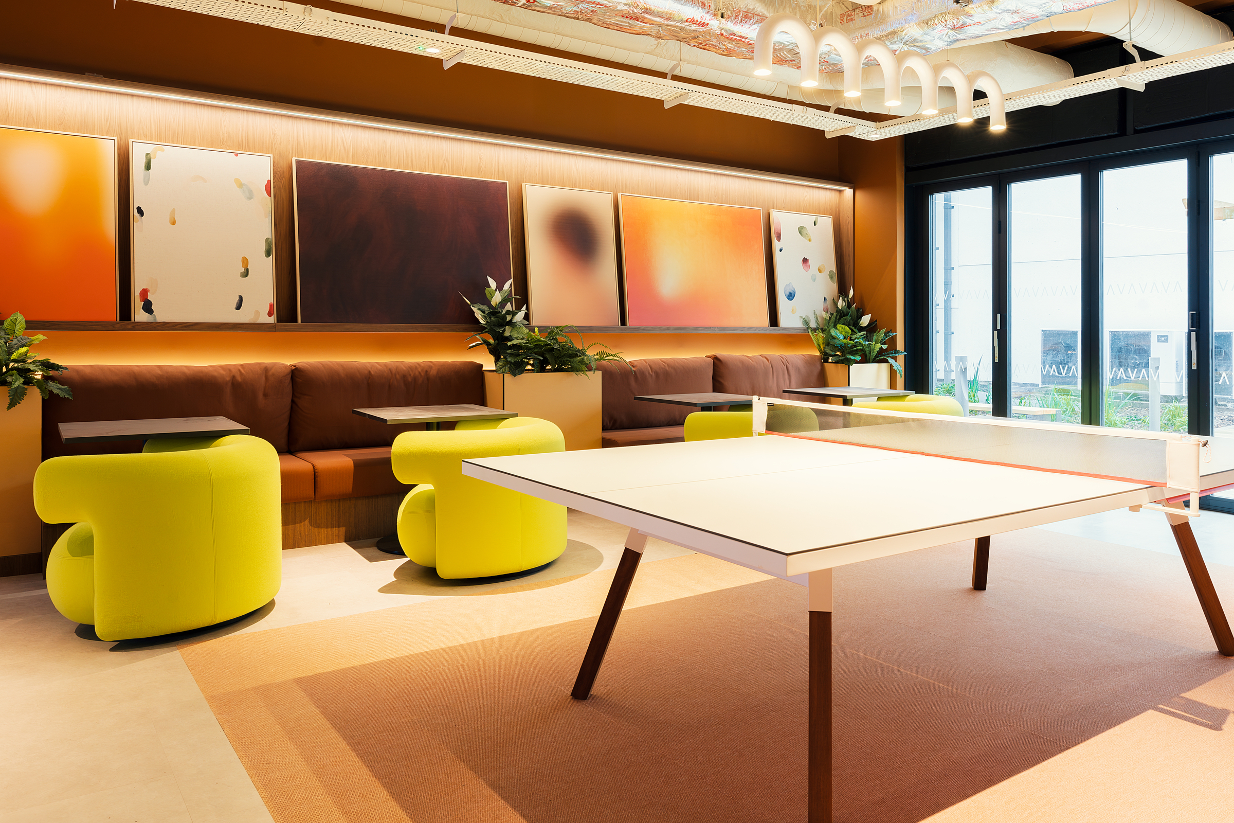 Modern lounge with yellow armchairs, brown sofas, a ping pong table, abstract art on the wall, and plants on tables. Large windows let in natural light, creating an inviting atmosphere in this stylish New Waverley setting.