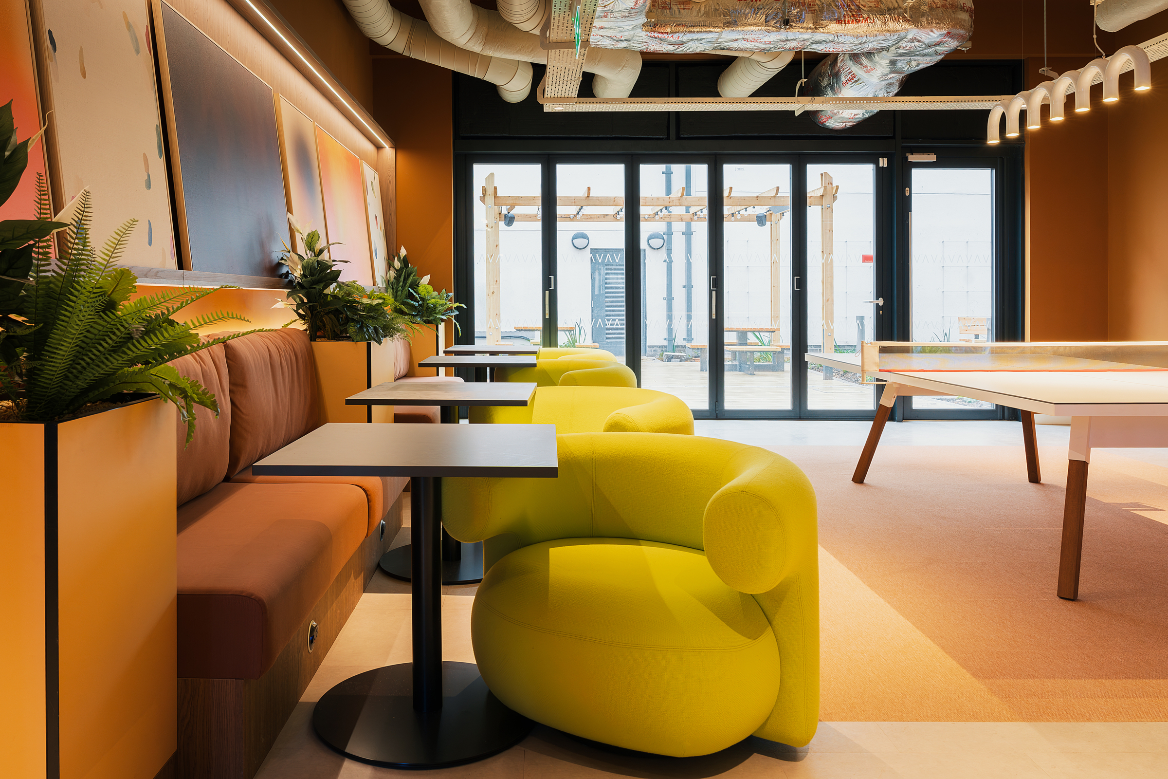 Modern lounge area with yellow armchairs, brown cushioned bench seating, indoor plants, small tables, and large windows letting in natural light. Exposed ceiling ductwork and contemporary decor create a stylish atmosphere inspired by the vibrant energy of New Waverley.