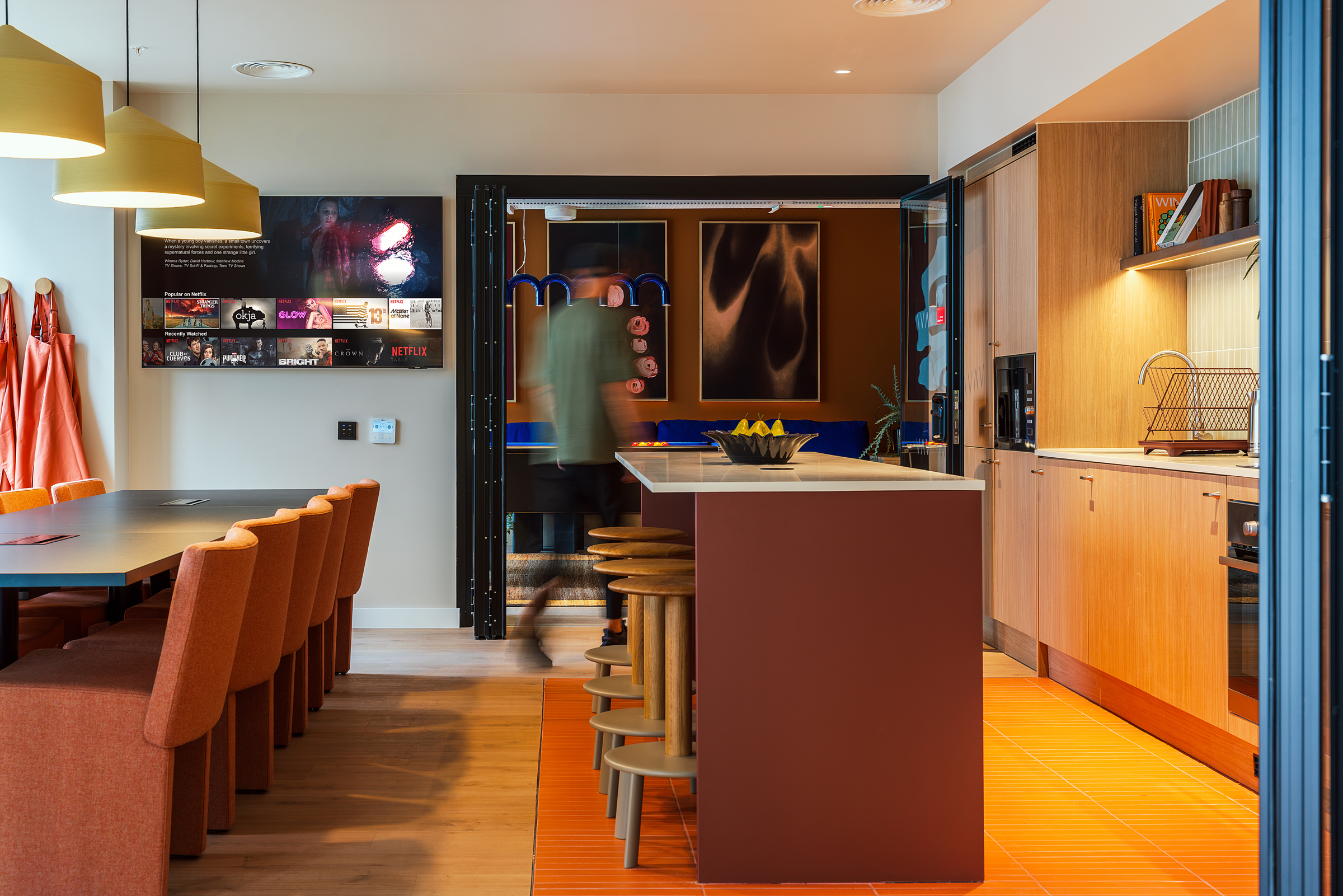 Modern communal kitchen and dining area at New Waverley with orange chairs, pendant lights, a large table, and a kitchen island. A blurred person walks toward an adjoining room featuring a blue pool table and vibrant wall art.
