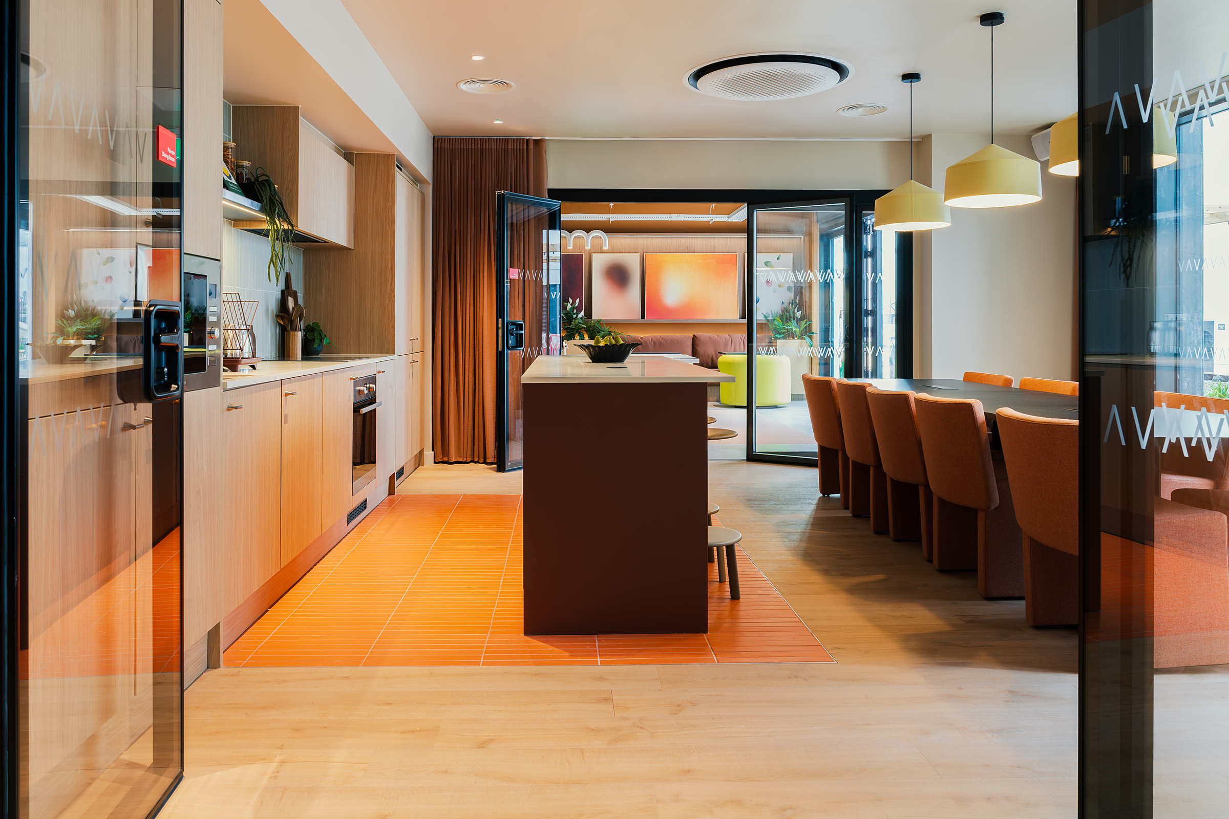 Modern kitchen and dining area with wooden cabinets, an island, orange chairs, hanging lights, and large windows. A lounge space with bright decor is visible through open glass doors in the background, creating a vibrant atmosphere characteristic of New Waverley living.