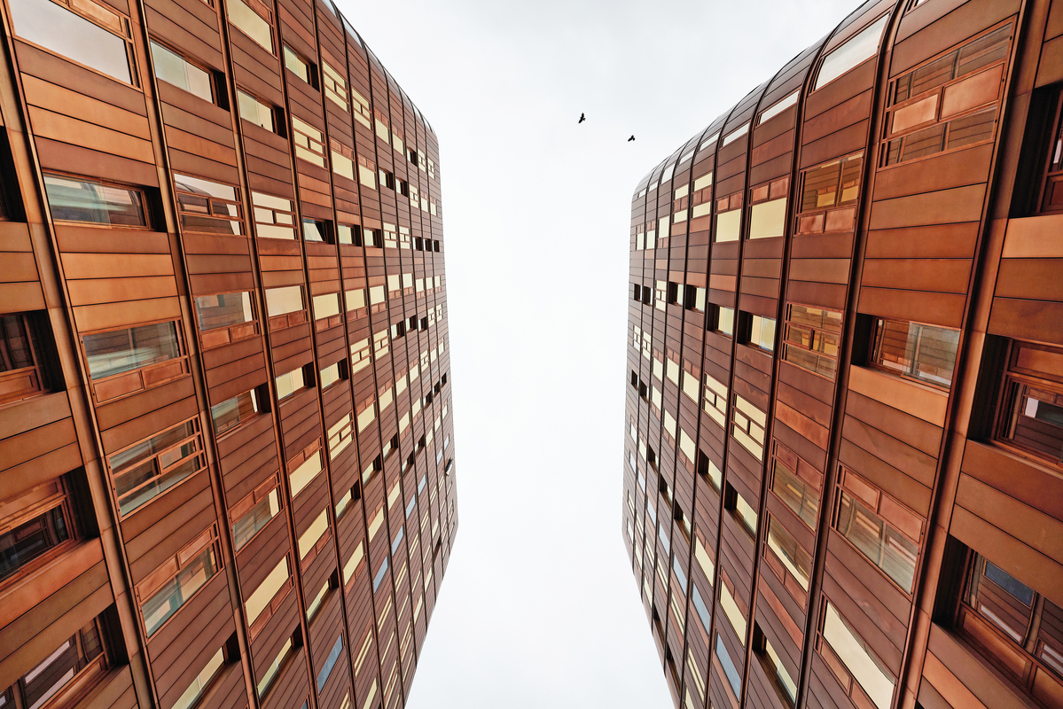 Two tall, modern buildings with reddish-brown panels rise on either side of the image in Coventry, converging toward a cloudy sky. Two birds are visible flying in the open space between the buildings.