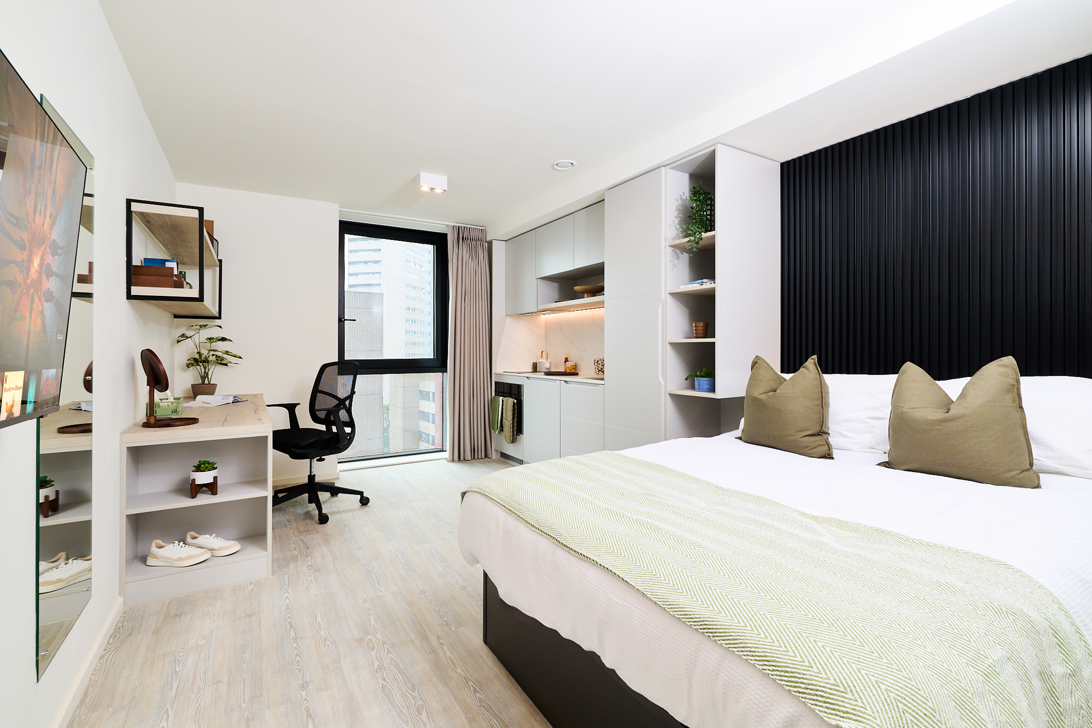 Modern studio apartment on Gough Street with a neatly made bed, small kitchenette, desk with chair, and large window showing city buildings. Minimalist decor with green accents and light wood flooring.
